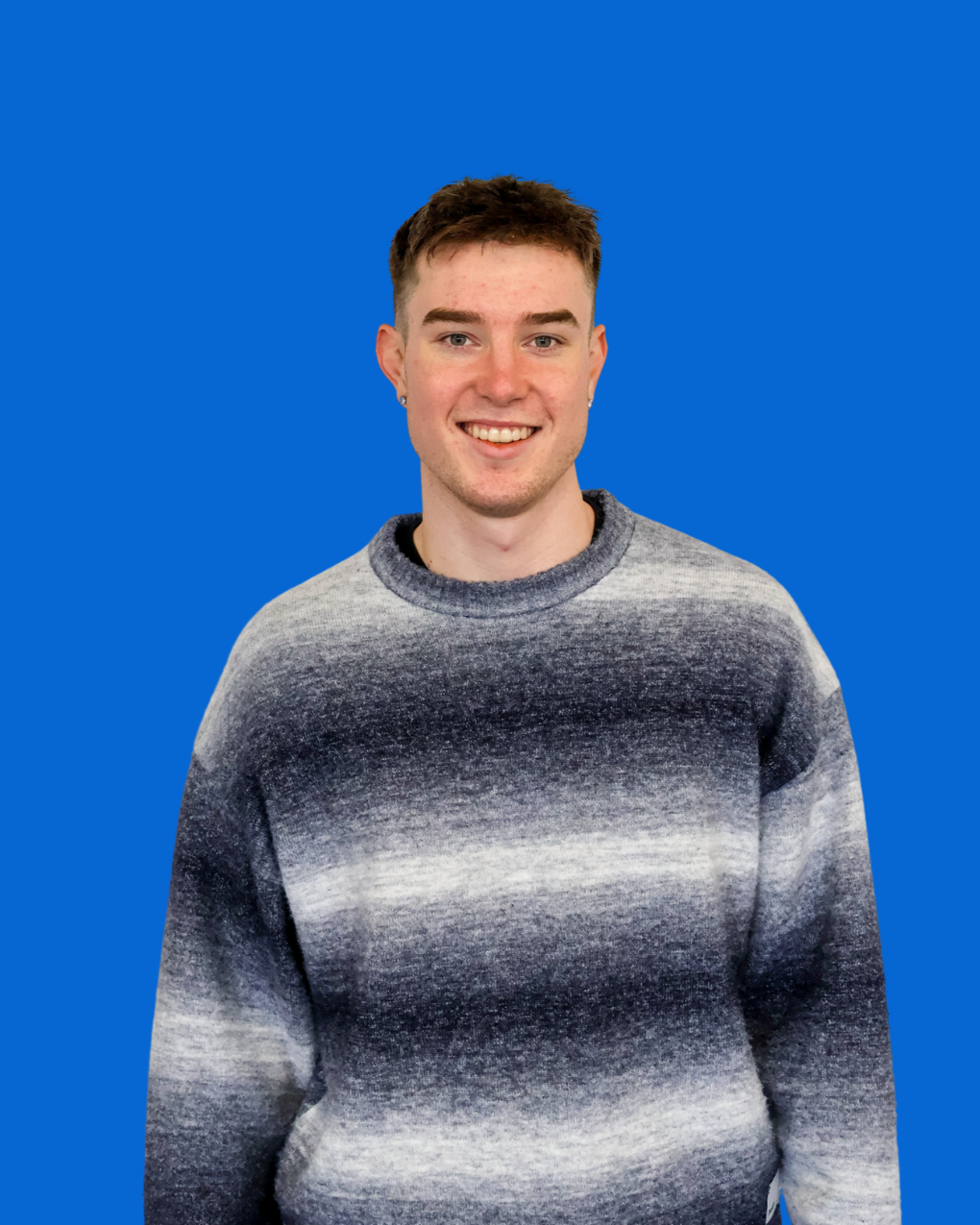 A smiling young man with short brown hair, wearing a gray knitted sweater with a gradient pattern, standing against a solid blue background.
