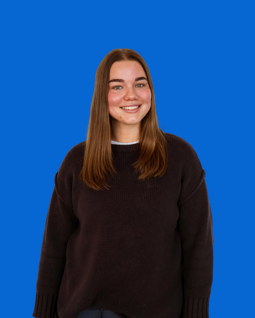 A young woman with long brown hair smiling, wearing a dark sweater, standing against a solid blue background.