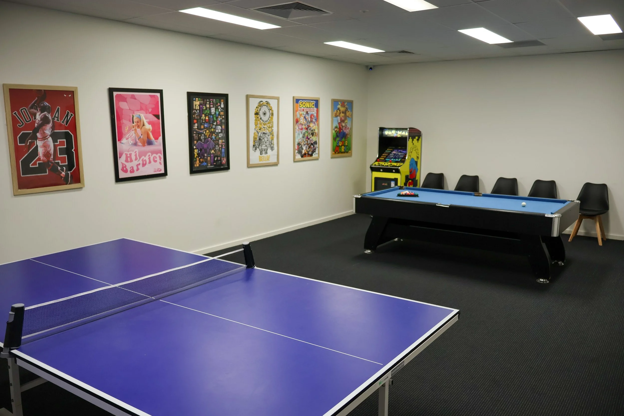 An indoor game room featuring a blue ping pong table, a basketball-themed poster, a pink Barbie poster, a pixel art poster, a Sonic the Hedgehog poster, a Pac-Man arcade game, and a row of black chairs along the wall. The room has a white ceiling with fluorescent lighting and a dark carpeted floor.