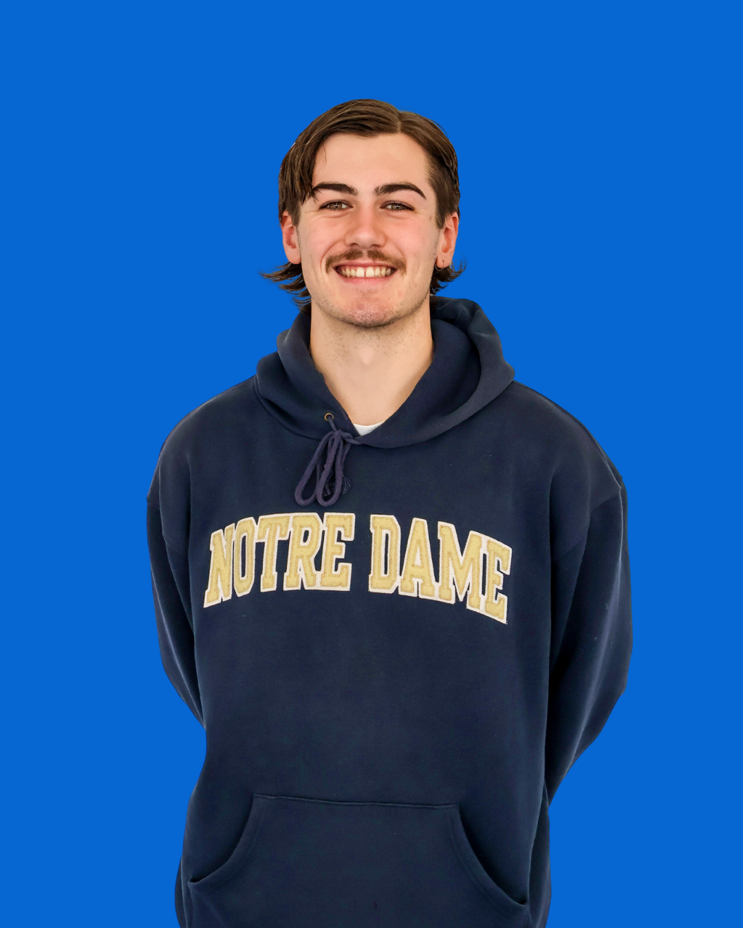 A young man with brown hair, mustache, and light skin, smiling, wearing a navy blue Notre Dame hoodie, standing against a solid blue background.