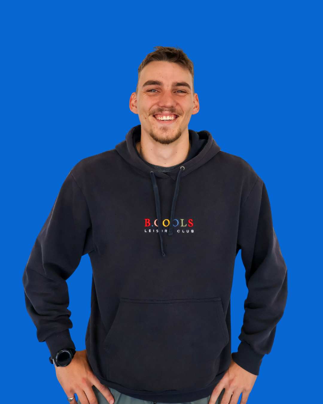 A young man with short brown hair, a light beard, and fair skin, smiling and standing against a solid blue background. He is wearing a black hoodie with colorful lettering on the front that reads 'B. COOLS LEISURE CLUB' and has his right hand on his hip.