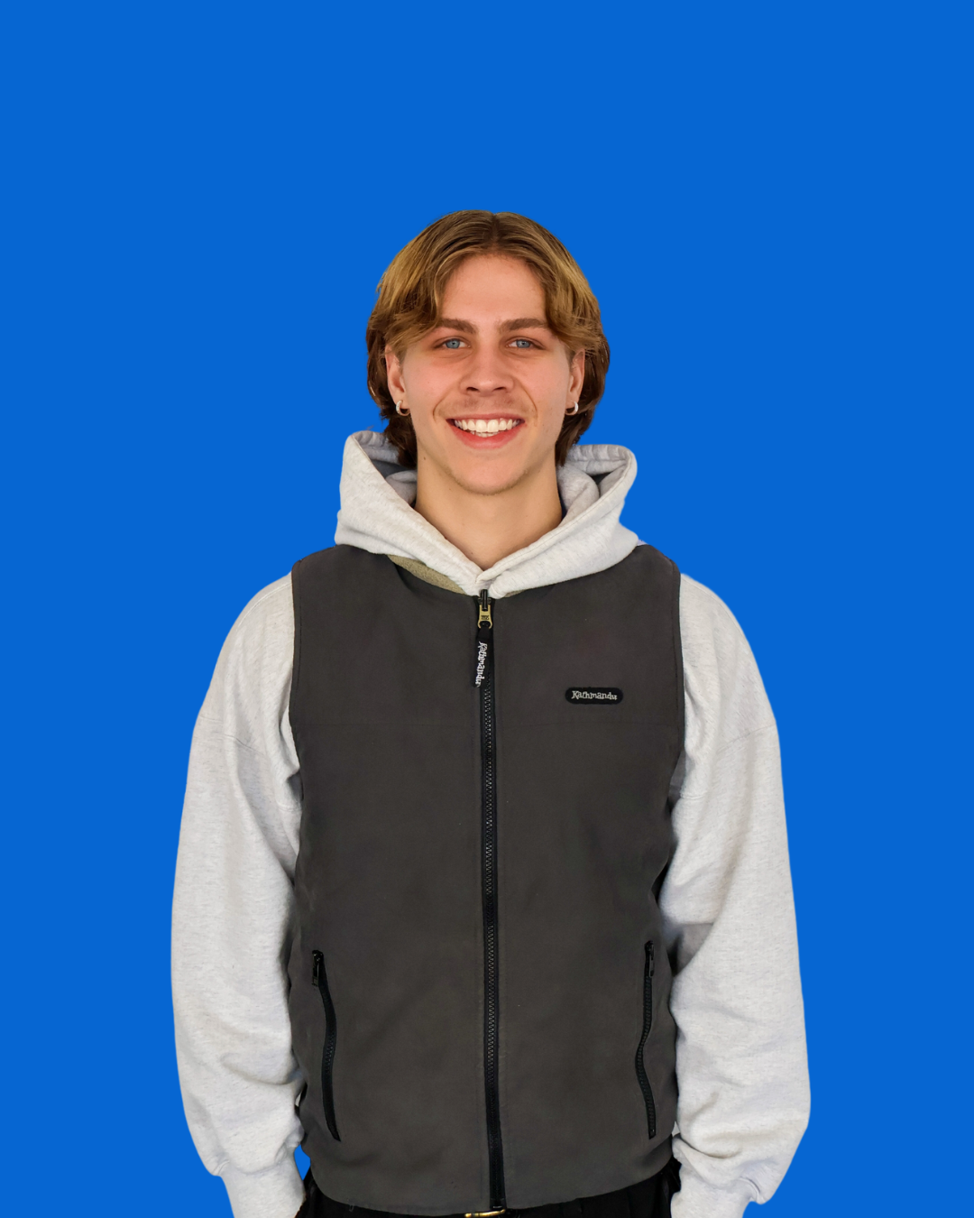 A young man with brown hair, smiling, wearing a gray hoodie and a black vest against a blue background.