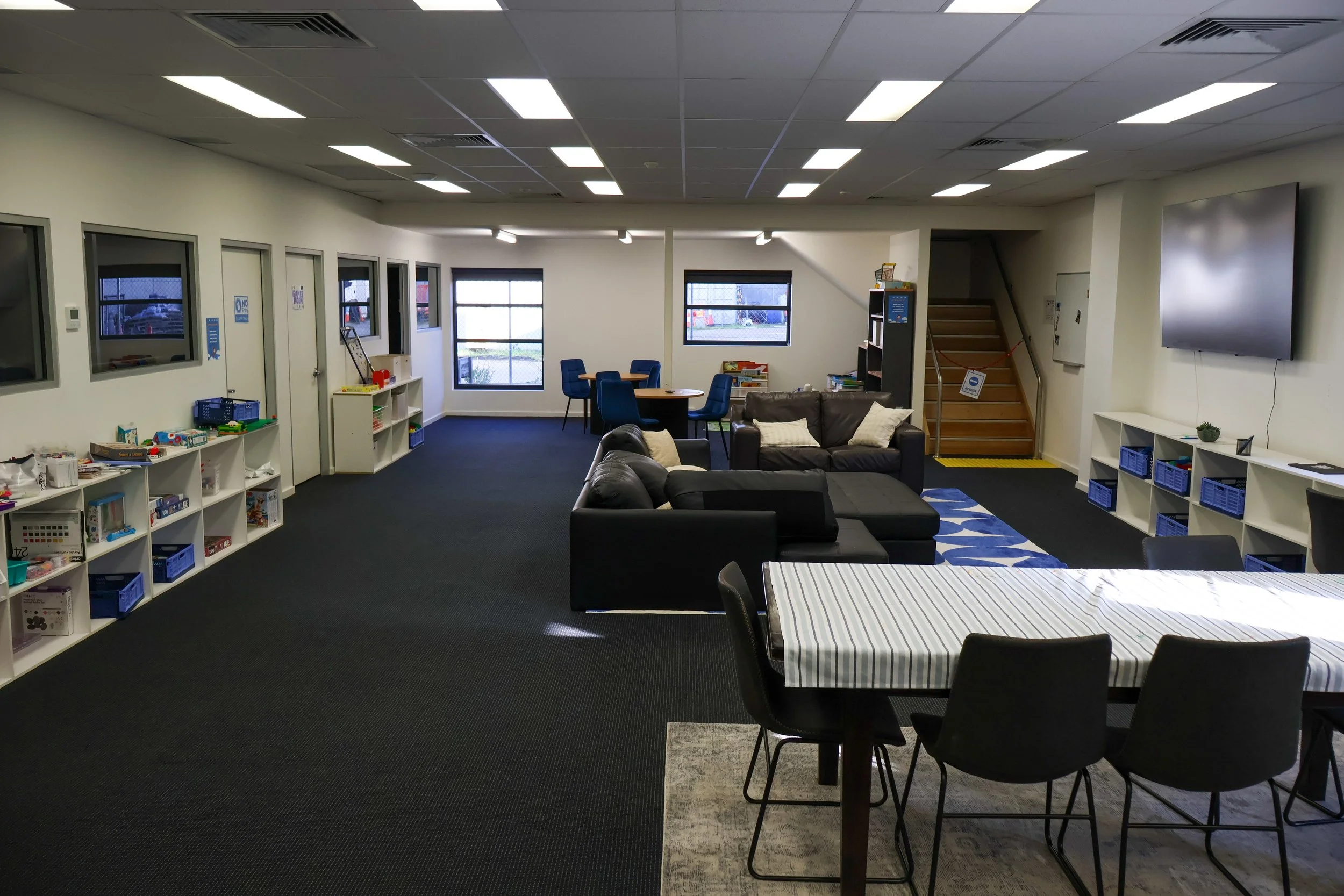 An indoor lounge area with black sofas, a table with a striped tablecloth, blue chairs around a small round table, white shelving units with books and toys, and a large wall-mounted TV. Windows reveal an outdoor view of parked cars.