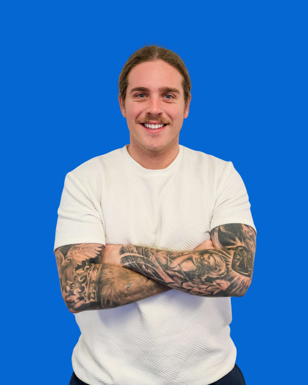 A young man with long brown hair, a mustache, and a nose piercing, smiling and standing with arms crossed against a solid blue background.