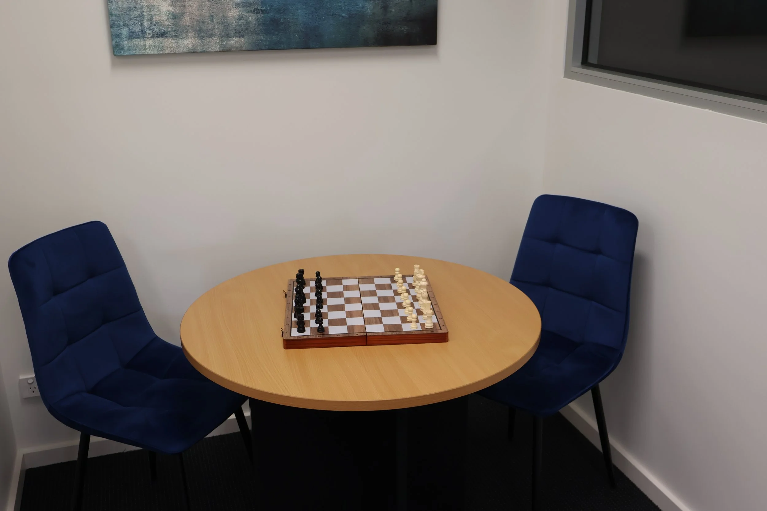 A round wooden table with a chessboard and chess pieces set up for a game, surrounded by three blue upholstered chairs in a modern office or conference room.