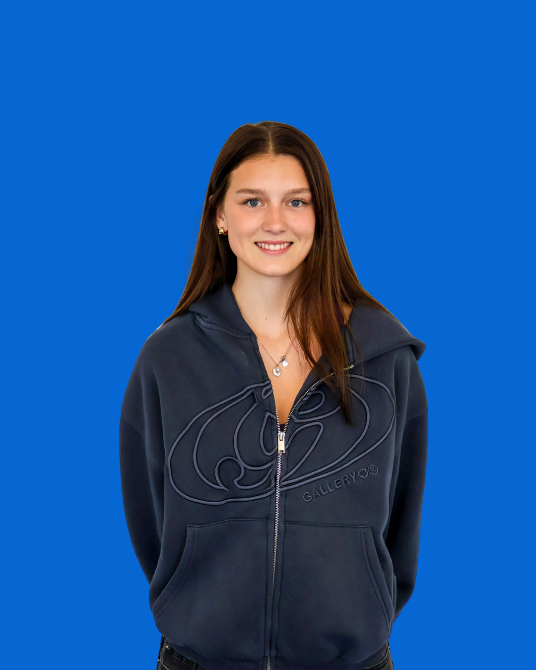 Young woman with long brown hair, wearing a dark zip-up hoodie with logo, standing against a bright blue background, smiling