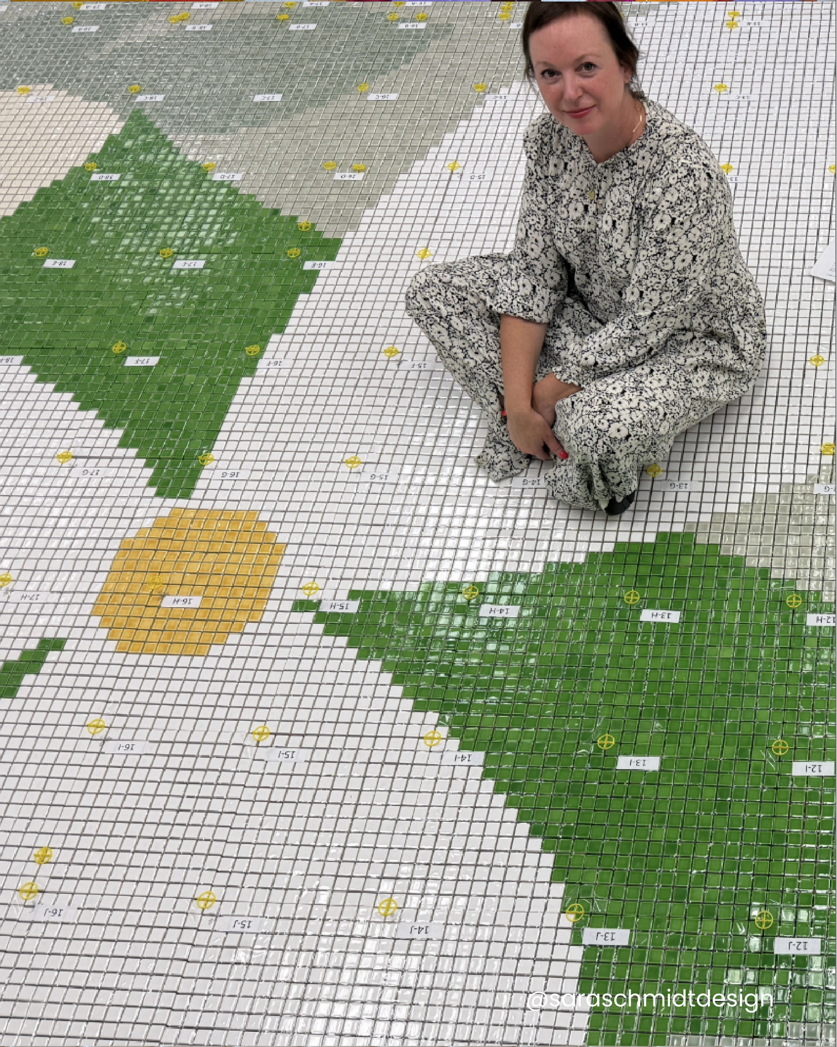 A woman in black and white patterned clothing sits on a floor covered in a large mosaic grid with colorful tiles forming a pattern, including green and yellow shapes.