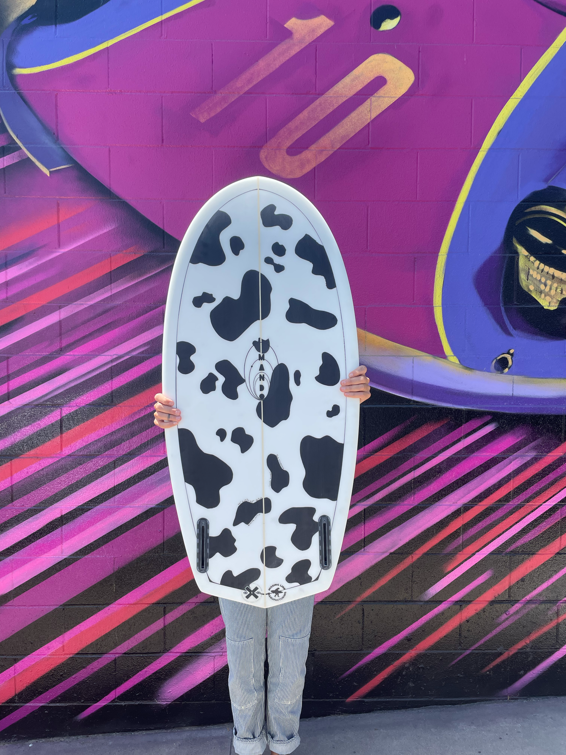 Person holding a cow print surfboard in front of a colorful, graffiti-style mural with abstract shapes and lines.