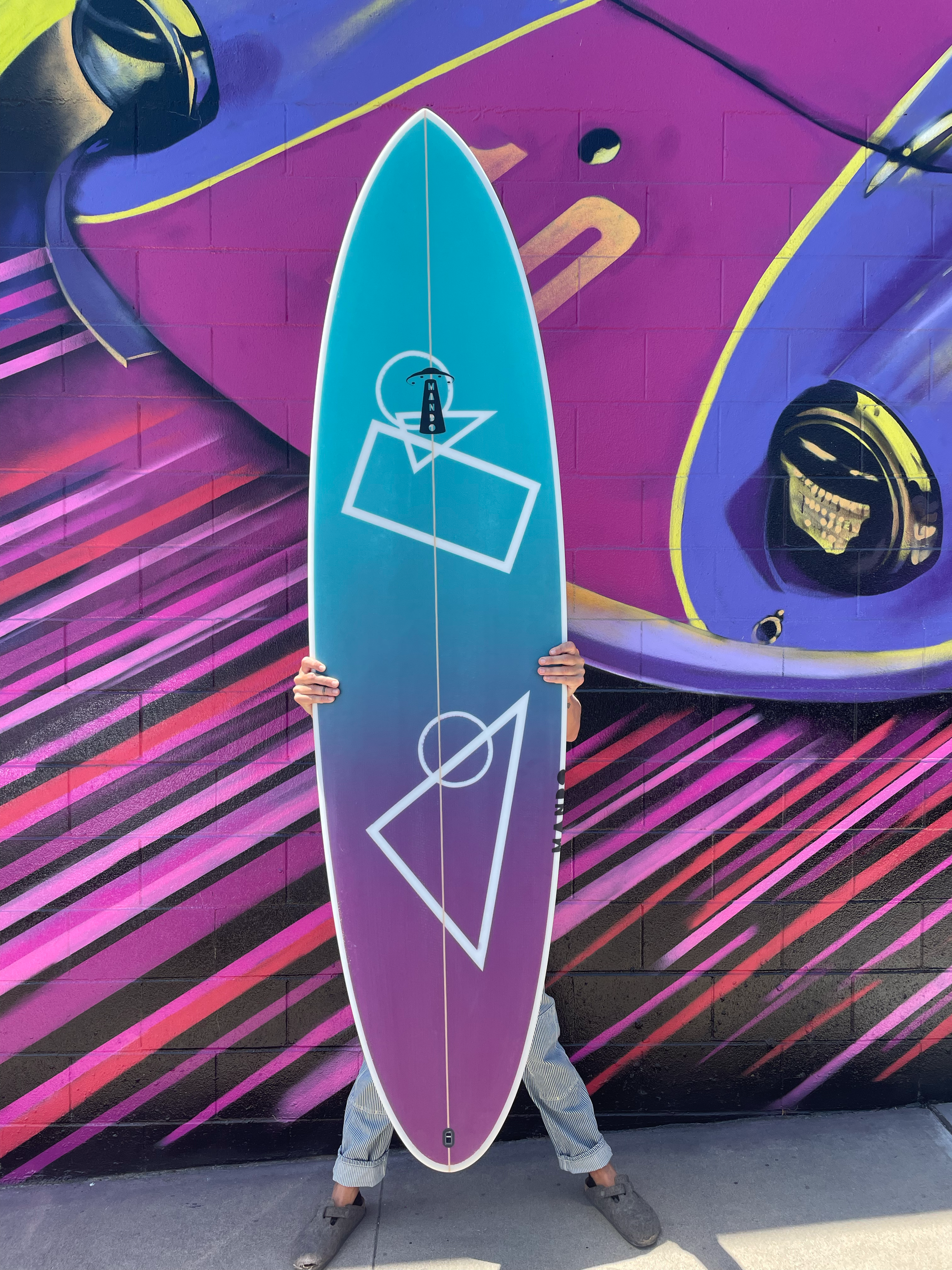 Person holding a colorful surfboard in front of a vibrant mural wall.