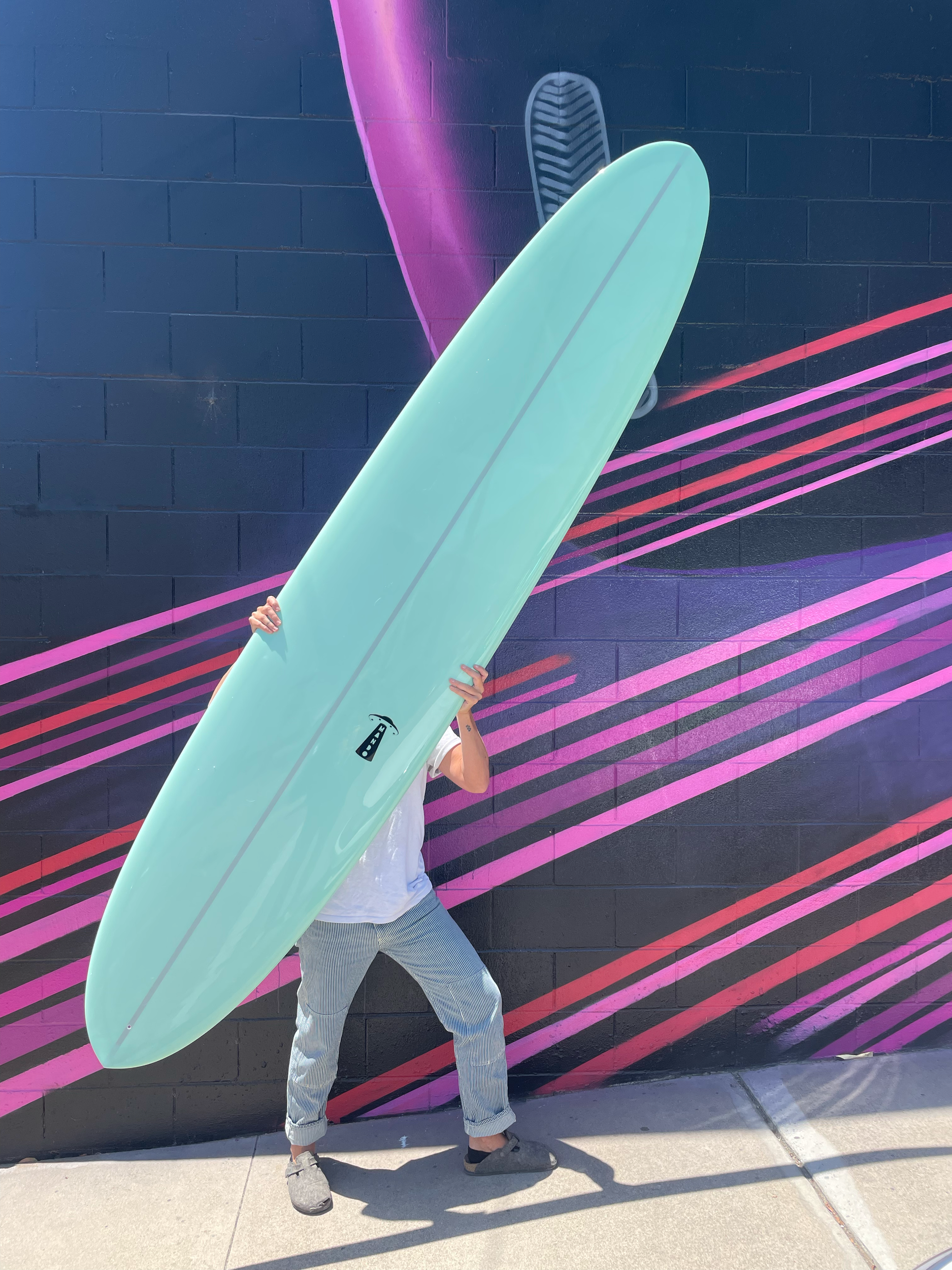 Person holding a large mint green surfboard in front of a colorful mural with pink and purple stripes and a black background.