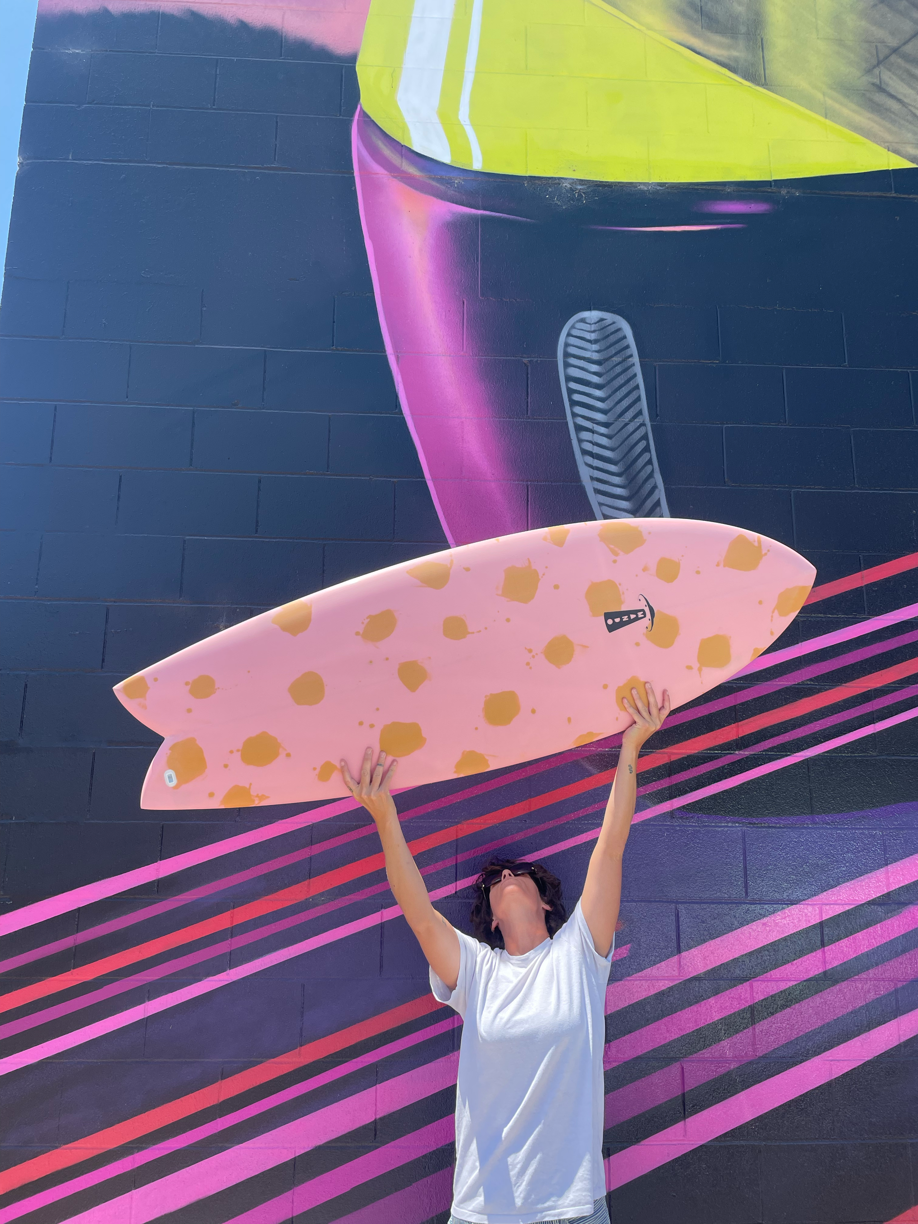 A person holding a pink surfboard with orange spots, standing against a colorful mural on a dark brick wall, with their arms raised and head tilted back.