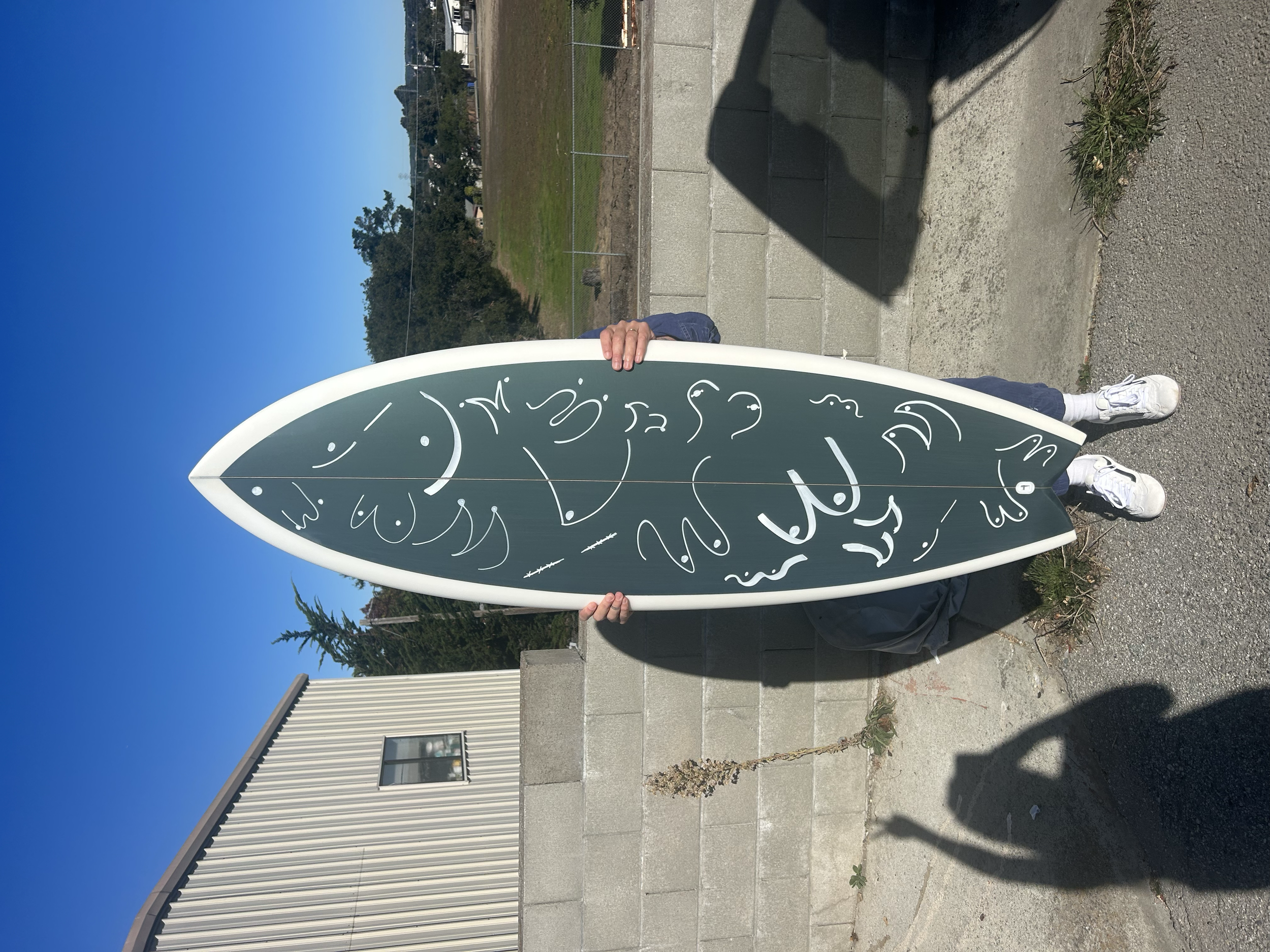 A person holding a black sandwich board sign with white decorative text outdoors on a sunny day. The person is wearing white sneakers and standing on a concrete sidewalk, with some small plants nearby. In the background, there is a grassy area with t