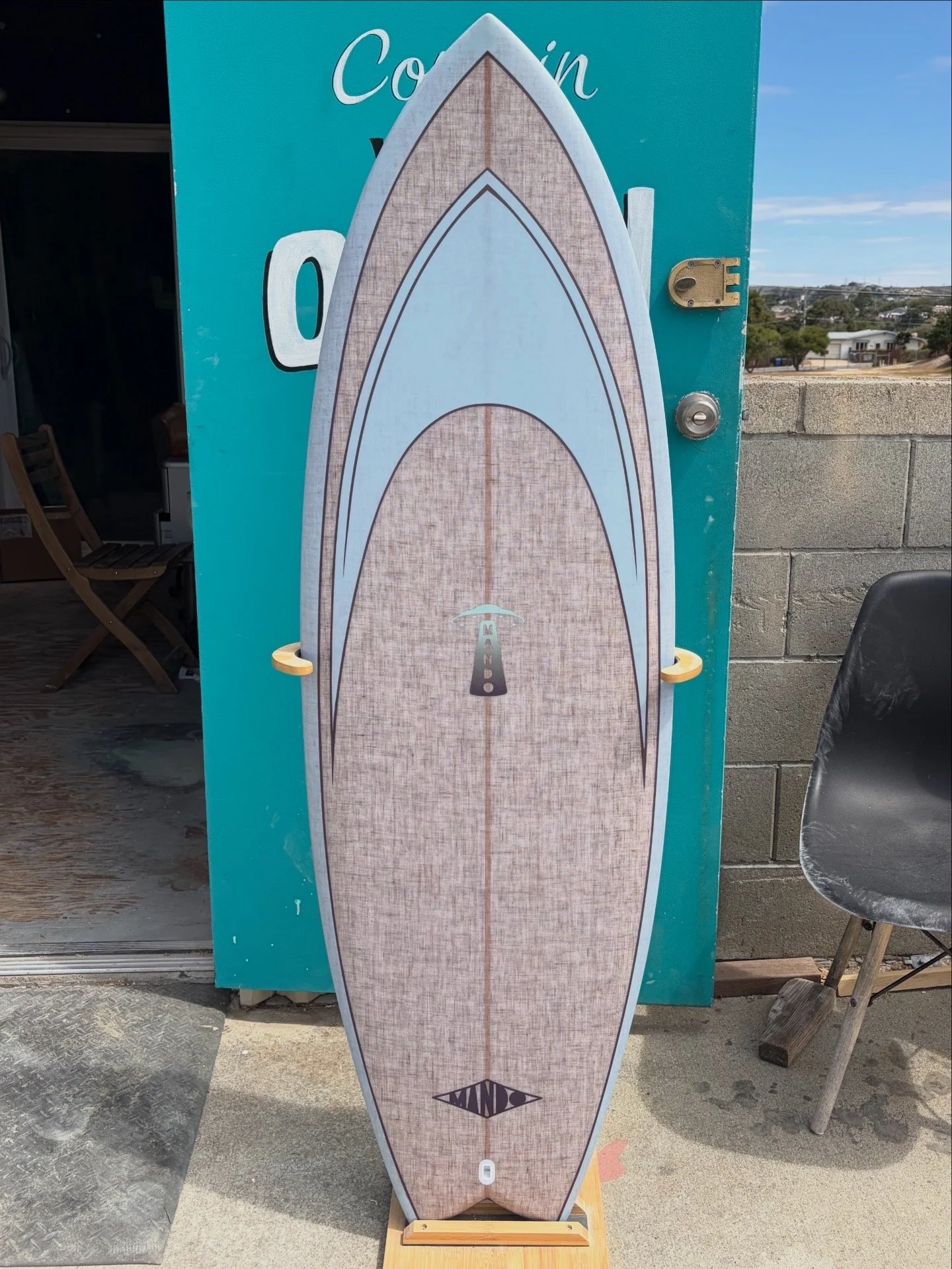 A surfboard with a wood grain pattern and a black diamond-shaped logo near the bottom, standing upright on a wooden stand outside a building, with a blue sign and a brick wall in the background.