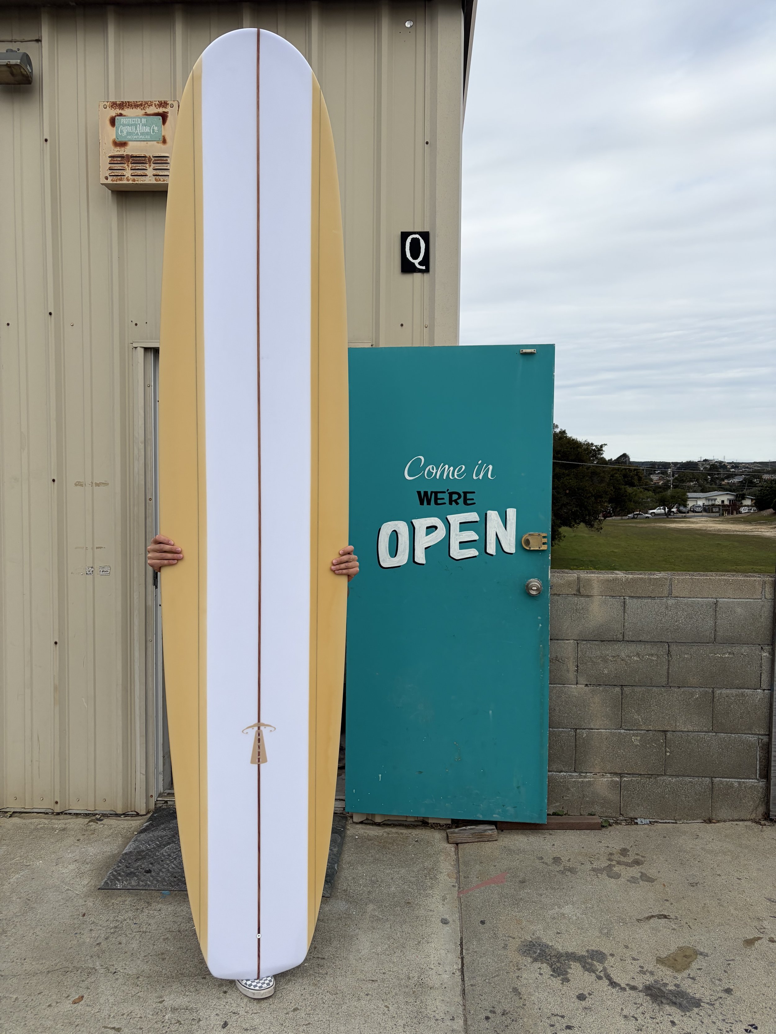 A person holding a long yellow surfboard with white and pastel yellow stripes standing outside a building with a teal 'Come in We're OPEN' sign and a black letter 'Q' sign on the wall, with a cloudy sky and distant horizon in the background.