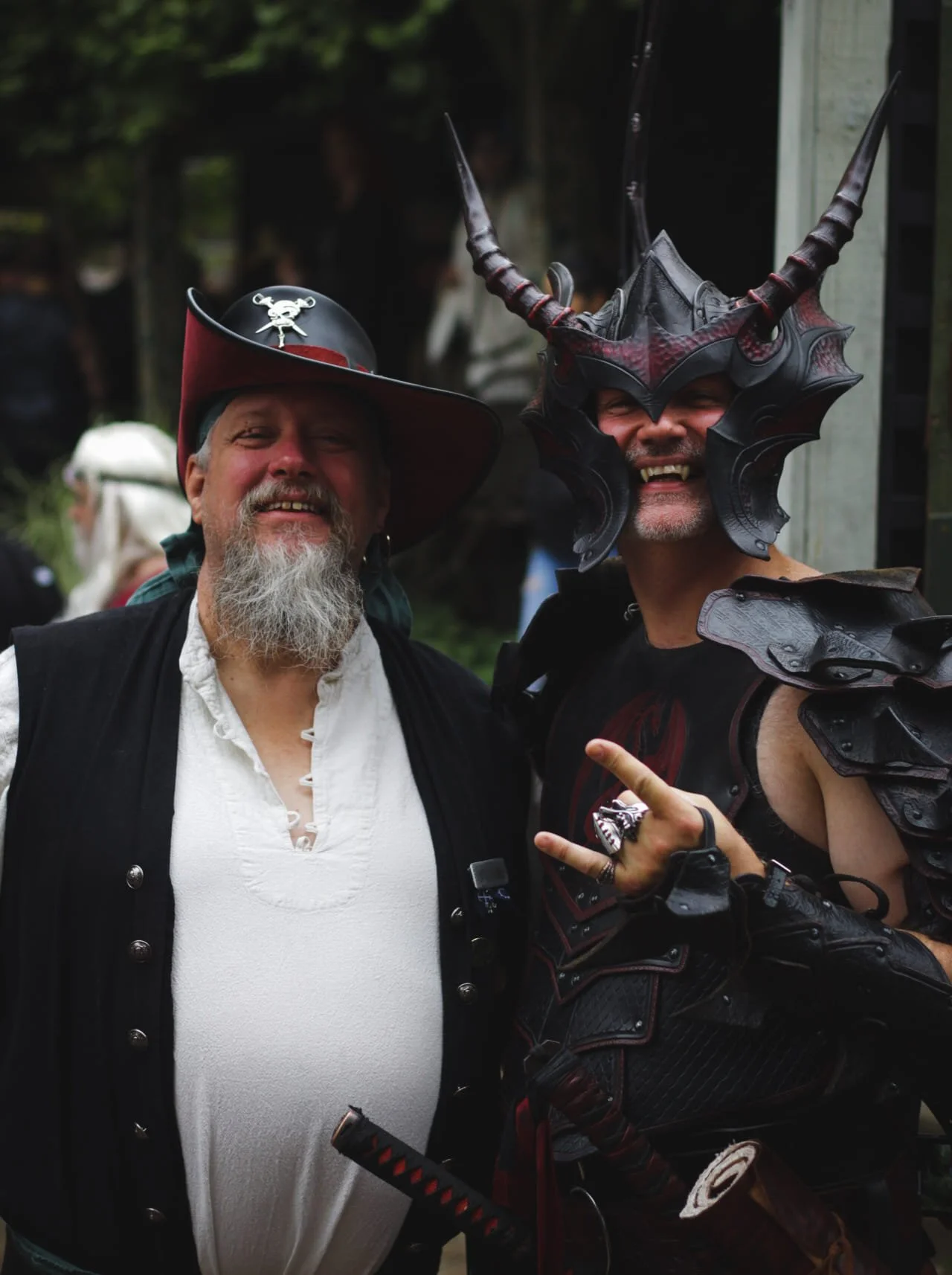 Two men dressed in fantasy and pirate costumes smiling at the camera, one wearing a pirate hat and the other a dragon-themed helmet, in an outdoor setting.