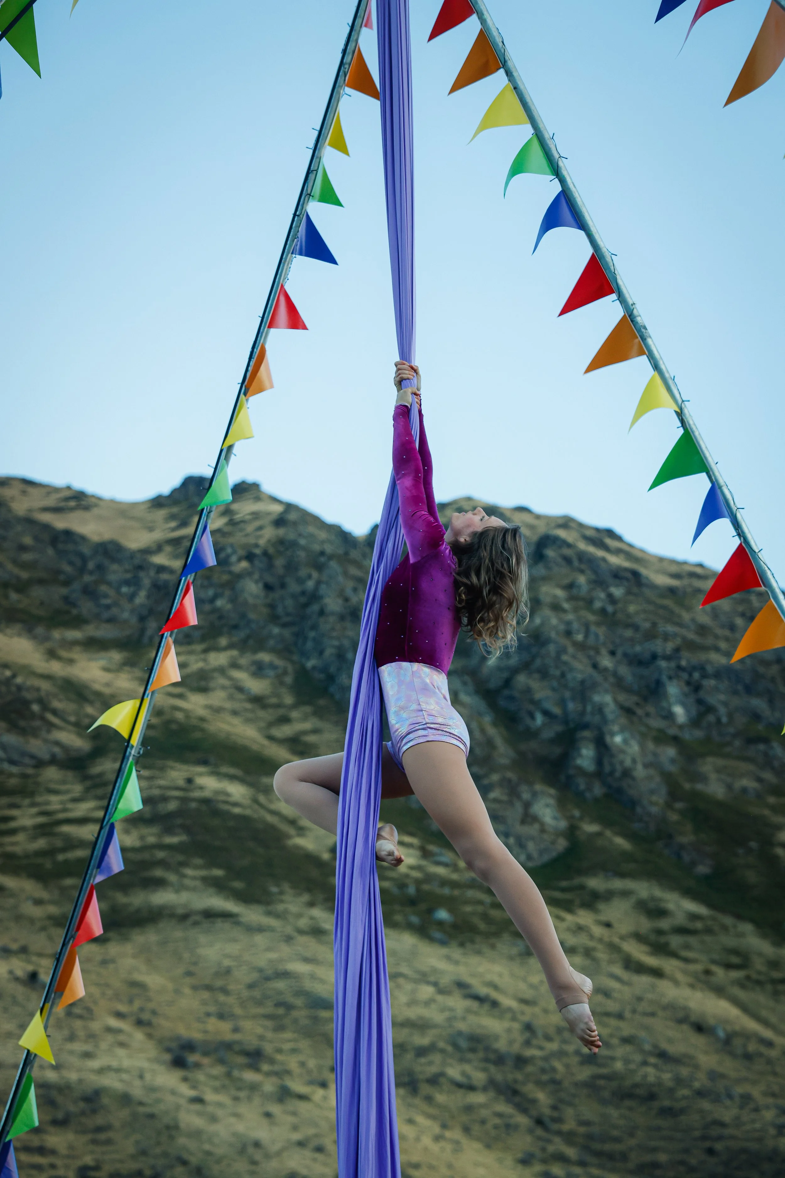 A young woman performs aerial silk acrobatics outdoors, hanging from purple silk with a mountain landscape in the background, surrounded by colorful flags.