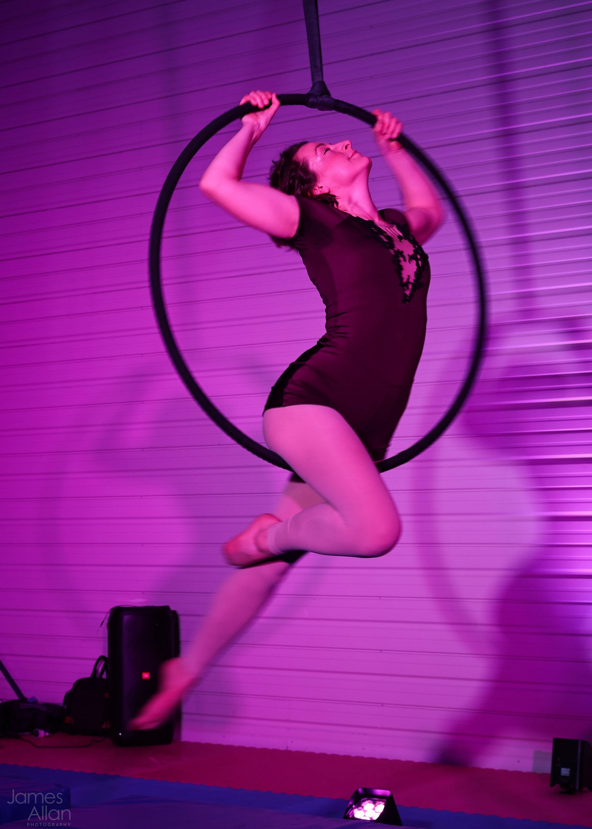 A woman performing on an aerial hoop in a pink-lit room, wearing a black leotard and tights.