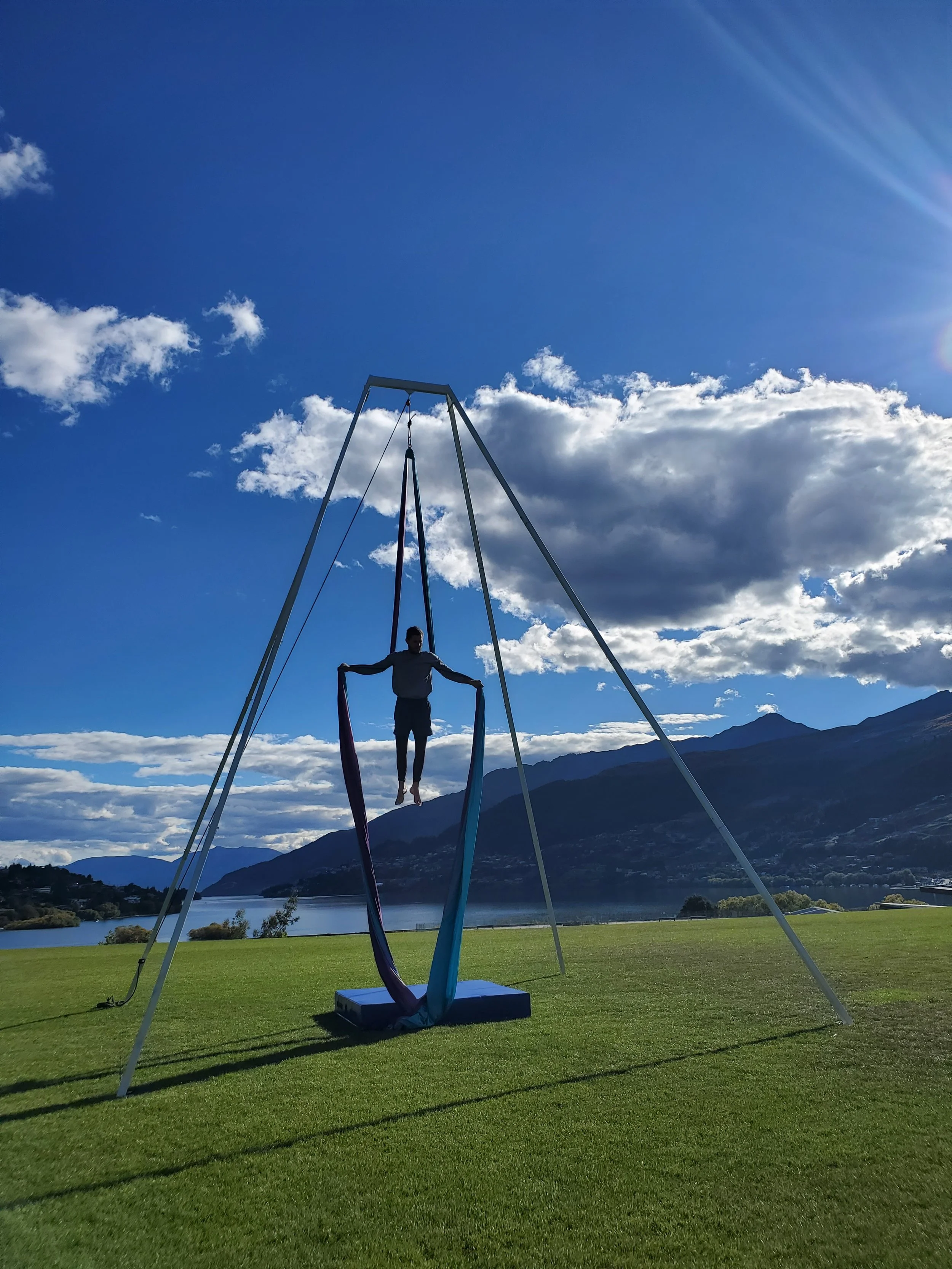 Person performing aerial silk acrobatics on a swing set outdoors with mountains and lake in the background.