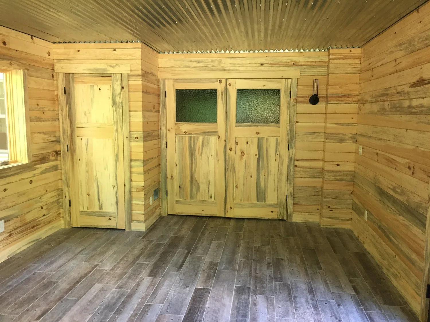 Interior of a room with wood-paneled walls, a wood door with a small window, and a ceiling with metal panels. The floor is covered with dark wooden planks.