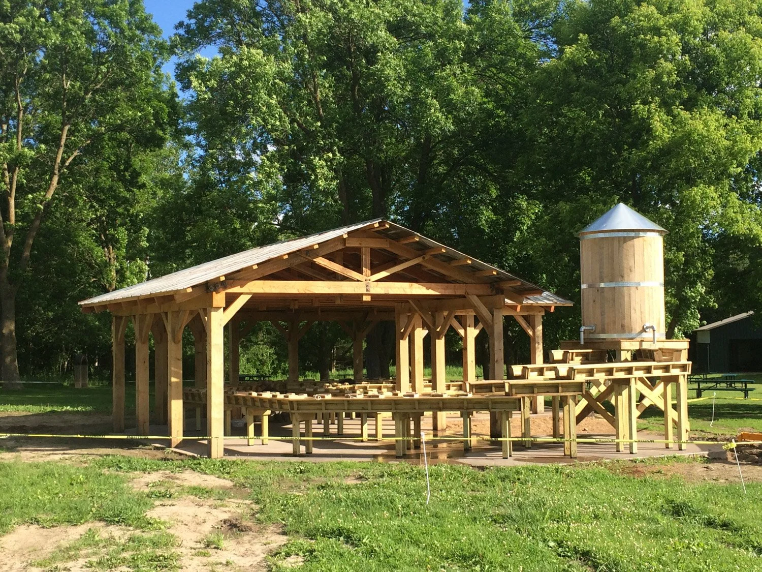 A wooden pavilion under construction with a raised wooden water tower nearby, set in a park with green trees in the background.