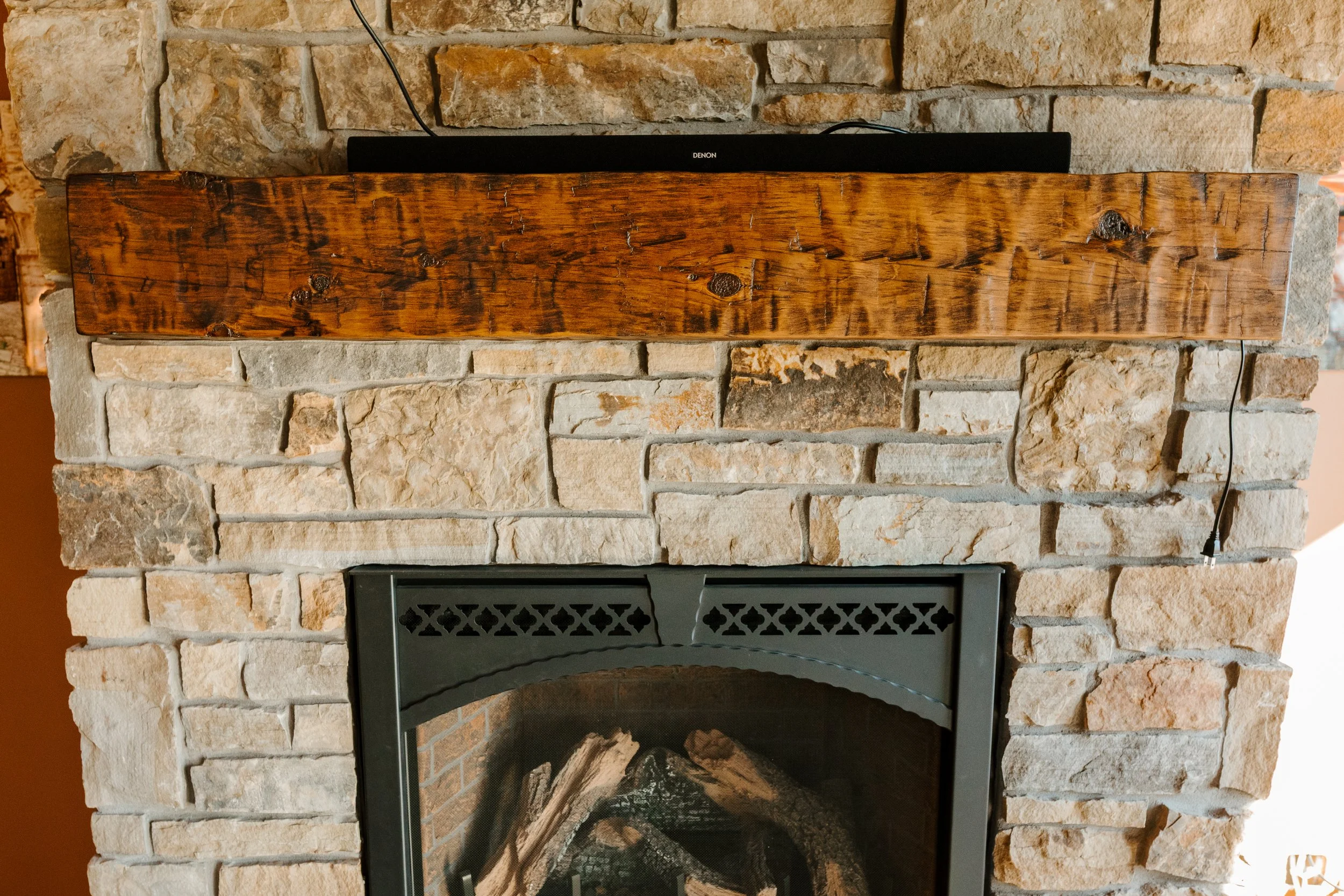 A stone fireplace with a rustic wooden mantle, with a black metal firebox opening at the bottom and a black soundbar on top of the mantle.