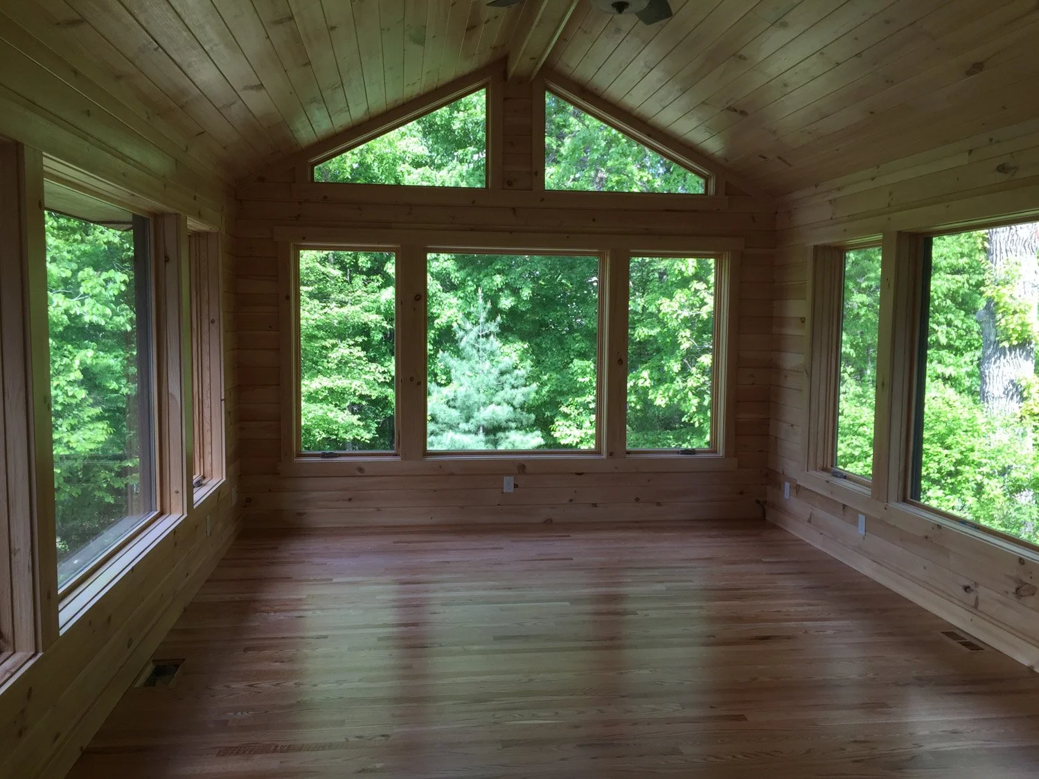 Empty room with large windows showing green trees outside, wooden walls, ceiling, and floor.
