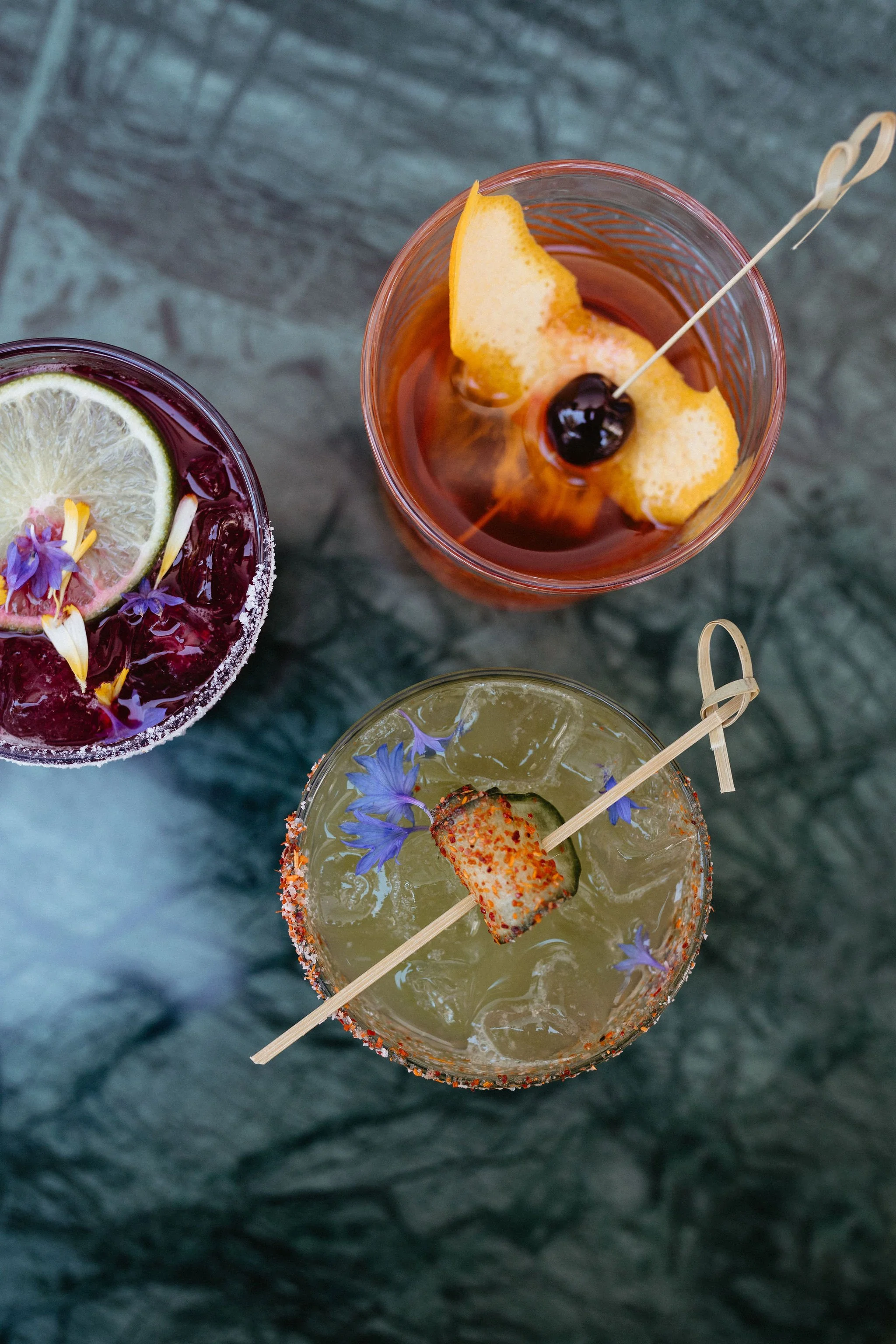 Three cocktails with garnishes on a dark table. The top right cocktail has an orange peel twist and a cherry skewer. The bottom cocktail has a slice of cucumber, edible purple flowers, and chili powder rim. The left drink has lemon and cherry garnish