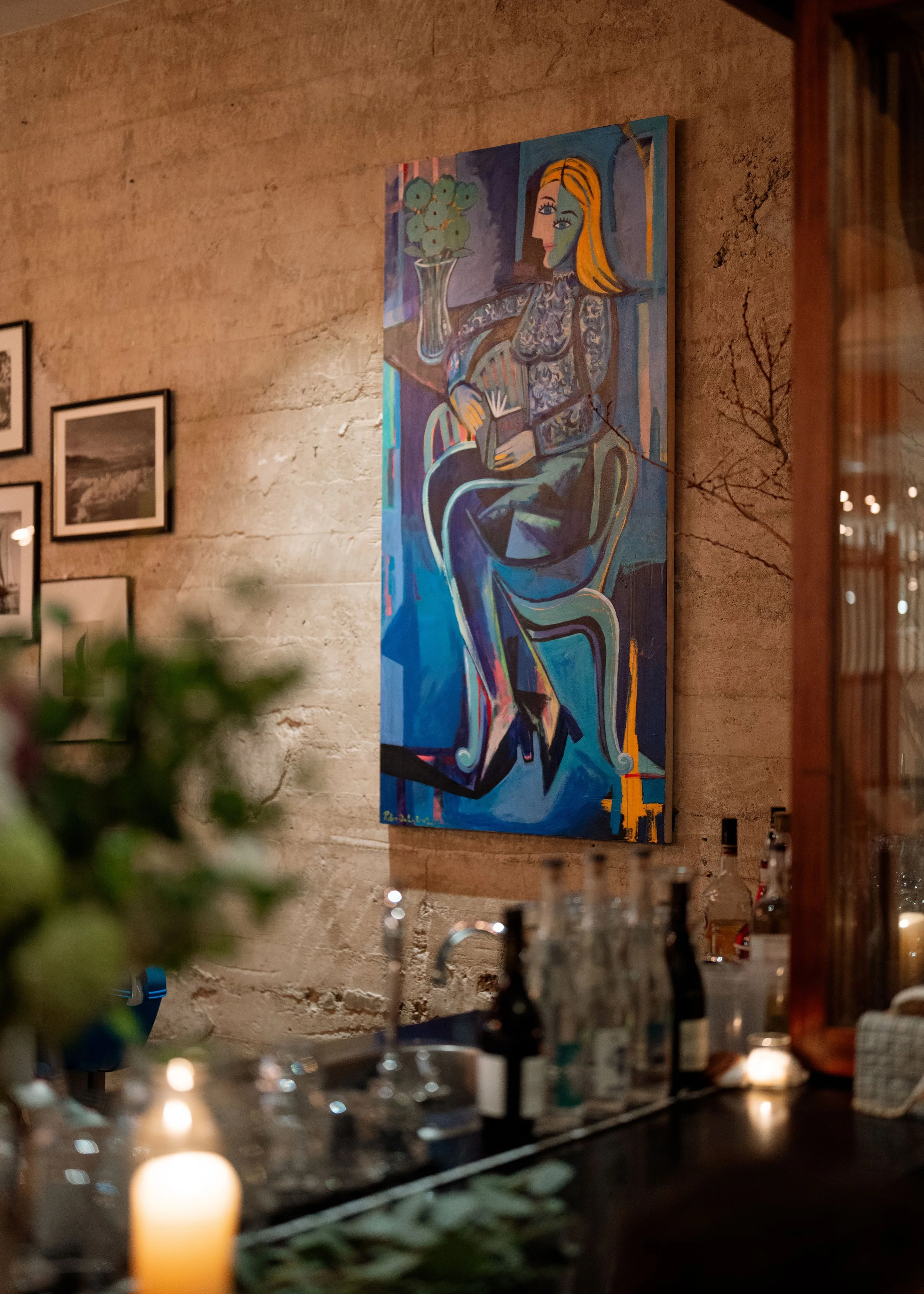 An interior view of a room with a stone wall and framed artwork, featuring a colorful painting of a woman with blonde hair sitting in a chair holding a book. The room has soft lighting, candles on a table, and some greenery in the foreground.