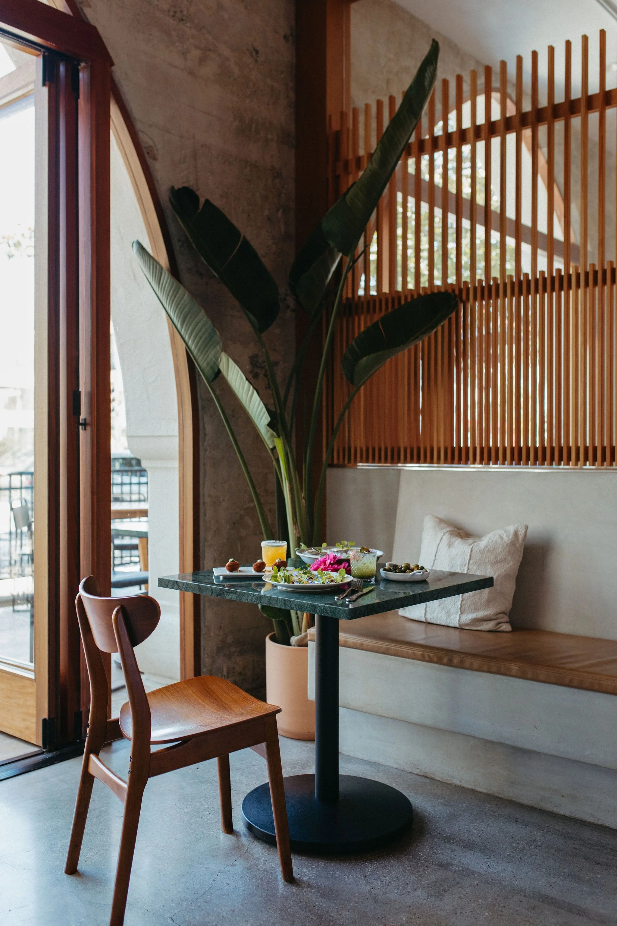 A cozy dining area with a small round table set with plates of food, drinks, and utensils. There is a wooden chair and a cushioned bench with a beige pillow. A large green plant with broad leaves stands next to a concrete wall. The space is lit by natural light coming through a large open window with wood framing, revealing an outdoor balcony with a table and chairs. A wooden slat room divider separates the dining space from the rest of the room.