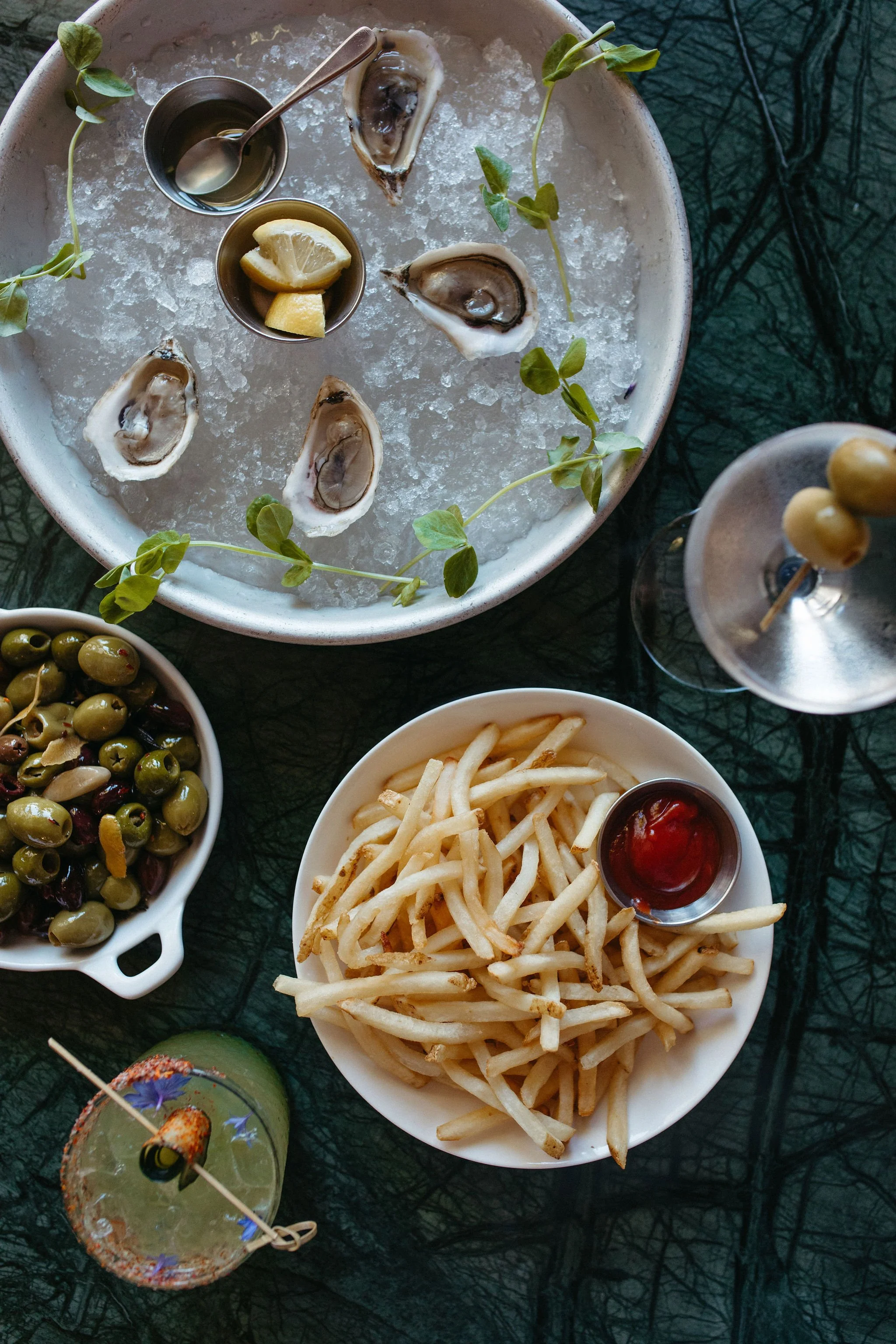 A table filled with various appetizers and drinks, including a bowl of green olives, a bowl of French fries with ketchup, a platter of oysters with lemon wedges, a small bowl of lemon slices, and a cocktail with a straw decorated with a flower, all o
