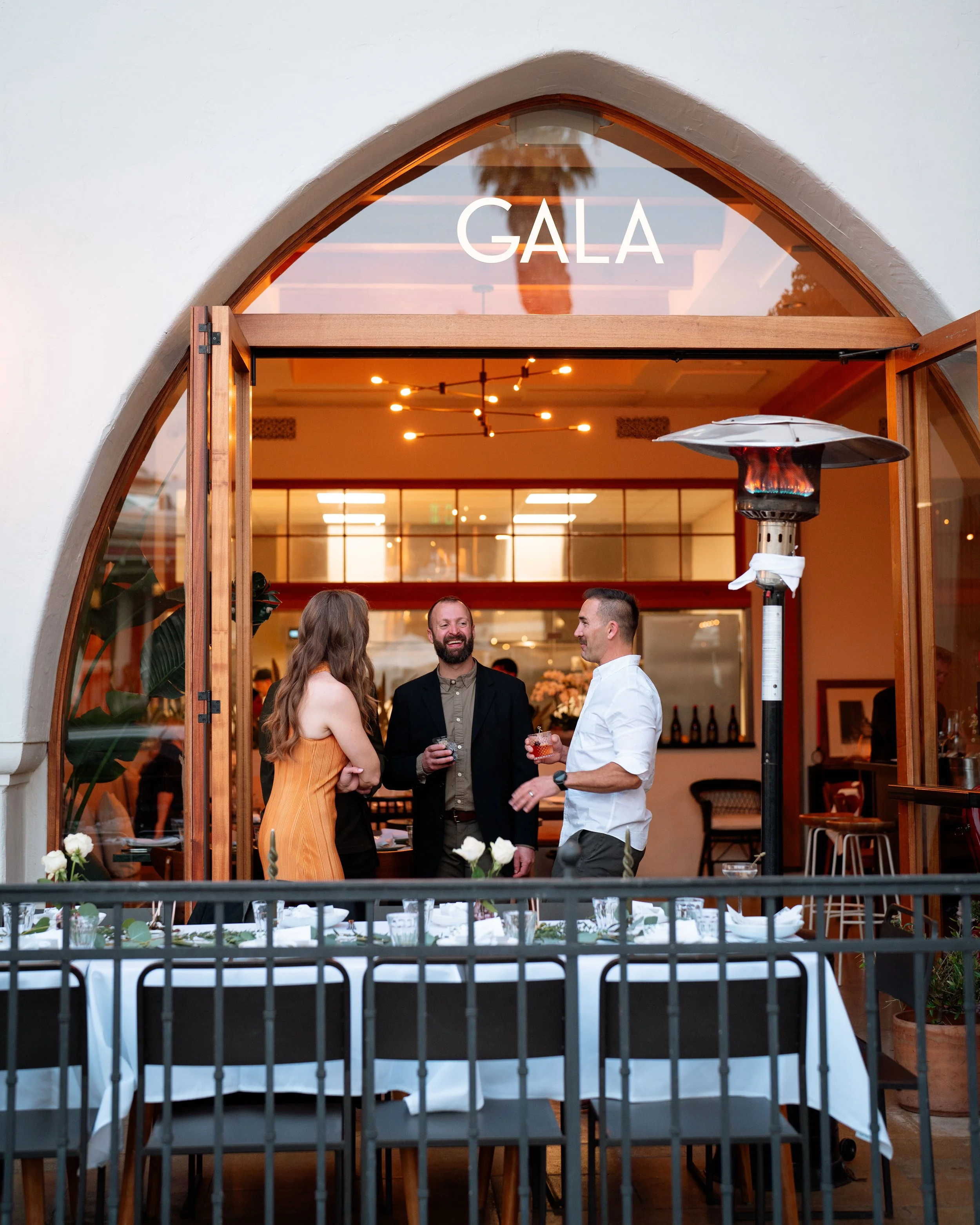 Indoor at a dinner event, people mingling and conversing near a patio heater, with a table set for a meal in the foreground, and a window with a sign reading 'GALA' above.