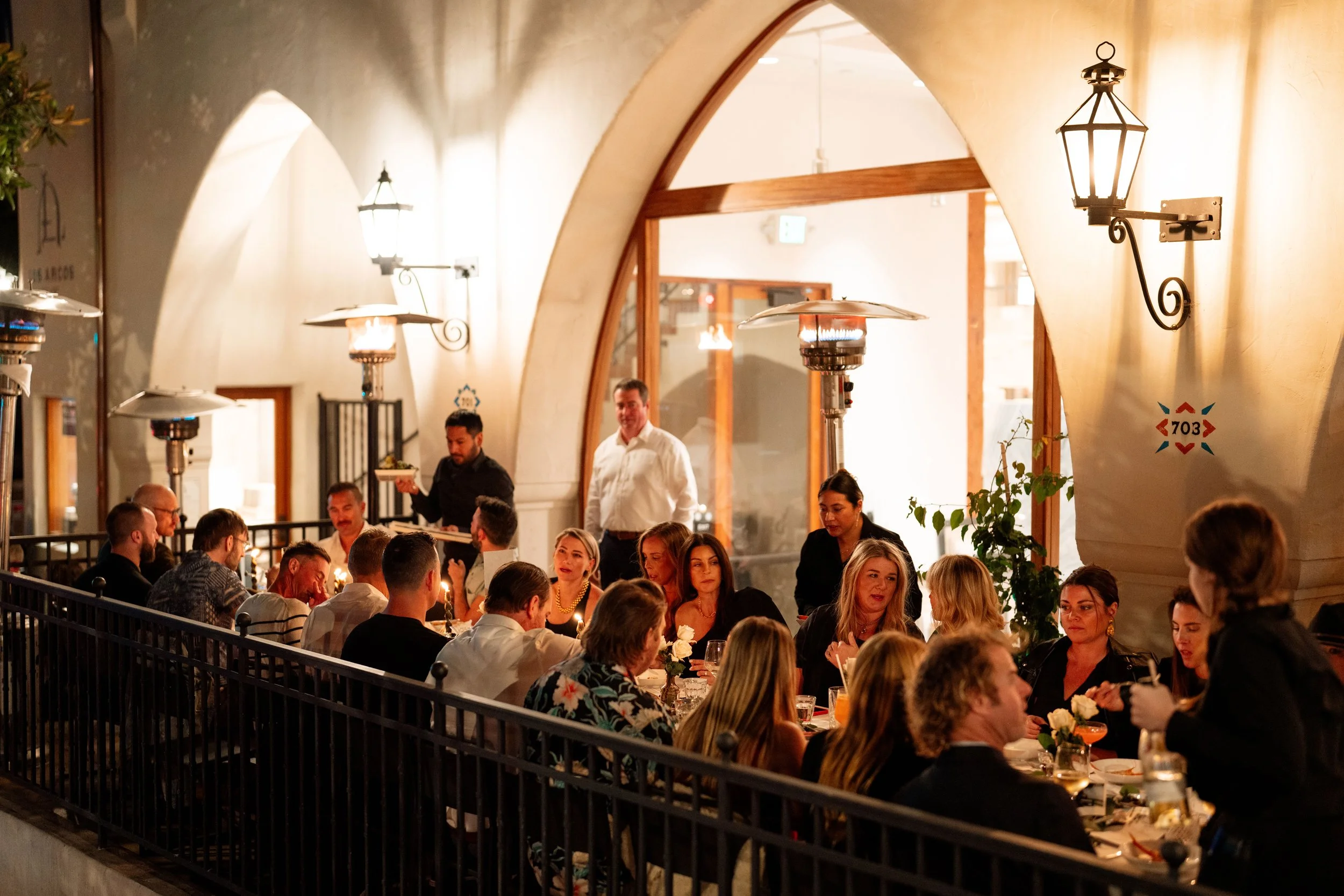 A group of people dining at a restaurant with dim lighting, decorated with elegant lantern-style wall sconces, situated under a curved ceiling. Two waiters are attending to guests at a long table.