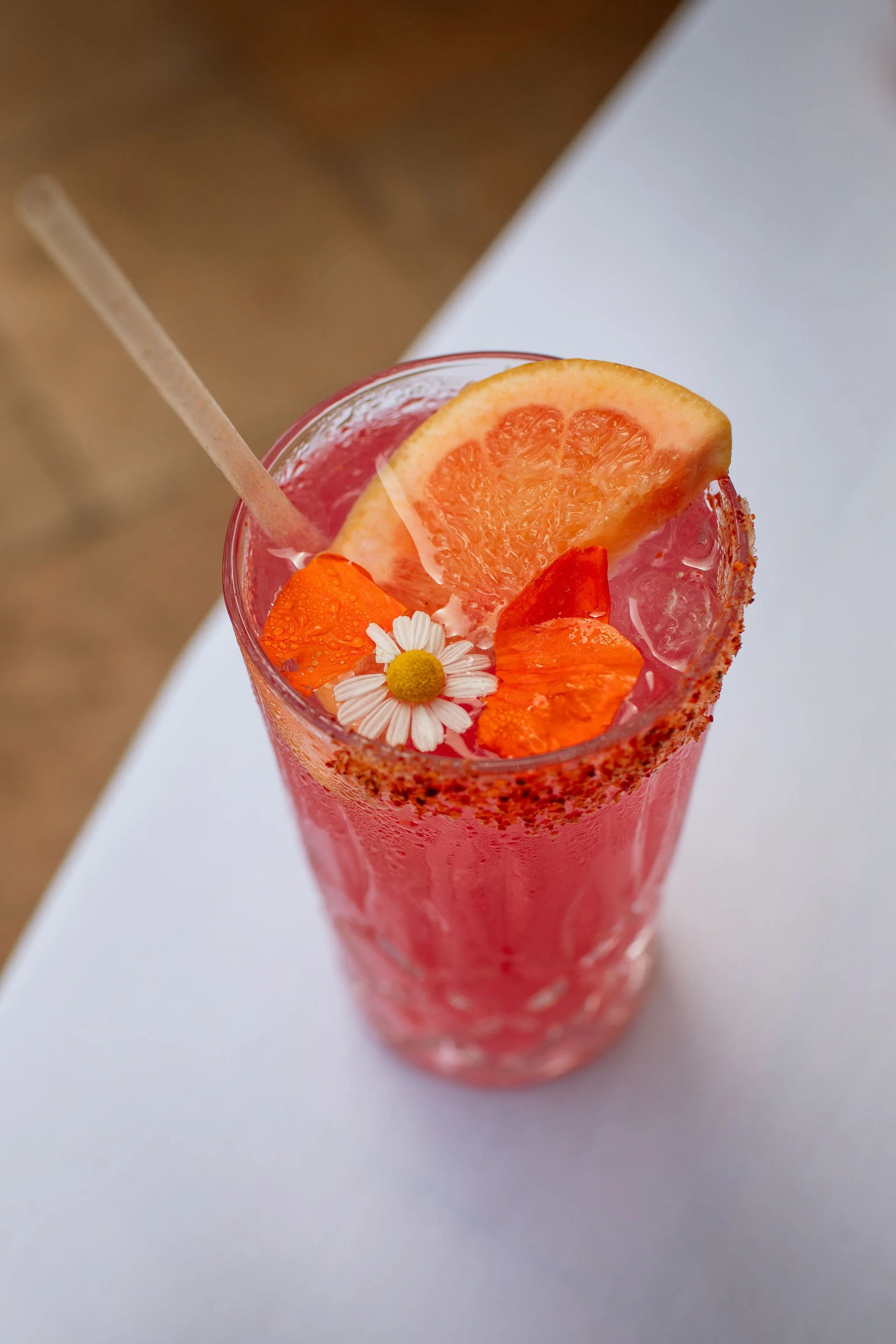 A tall glass of pink beverage garnished with a slice of grapefruit, an orange flower, and a white flower, with a salted rim and a straw.