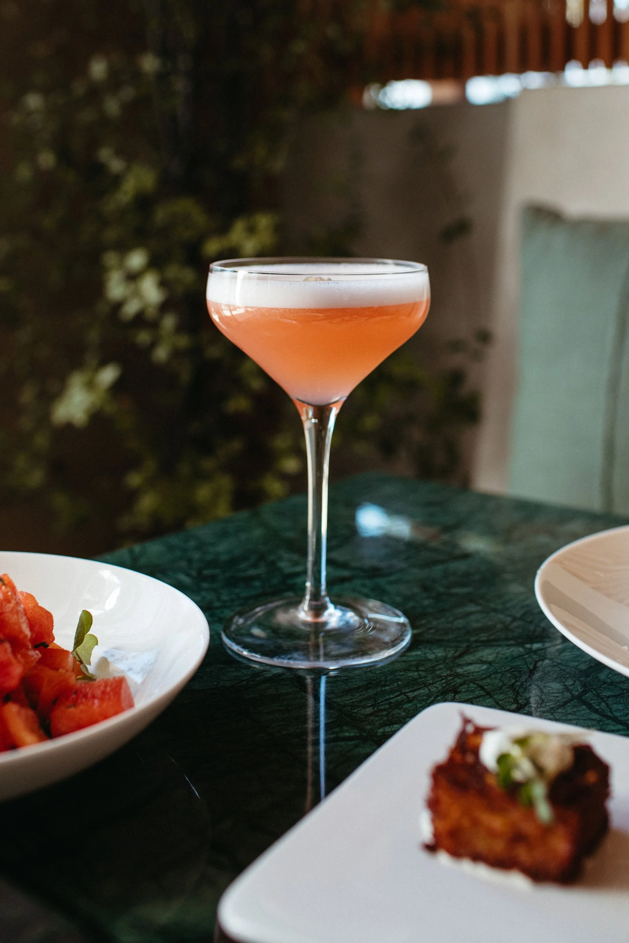 A pink cocktail in a coupe glass on a dark green marble table, with plates of food partially visible around it.