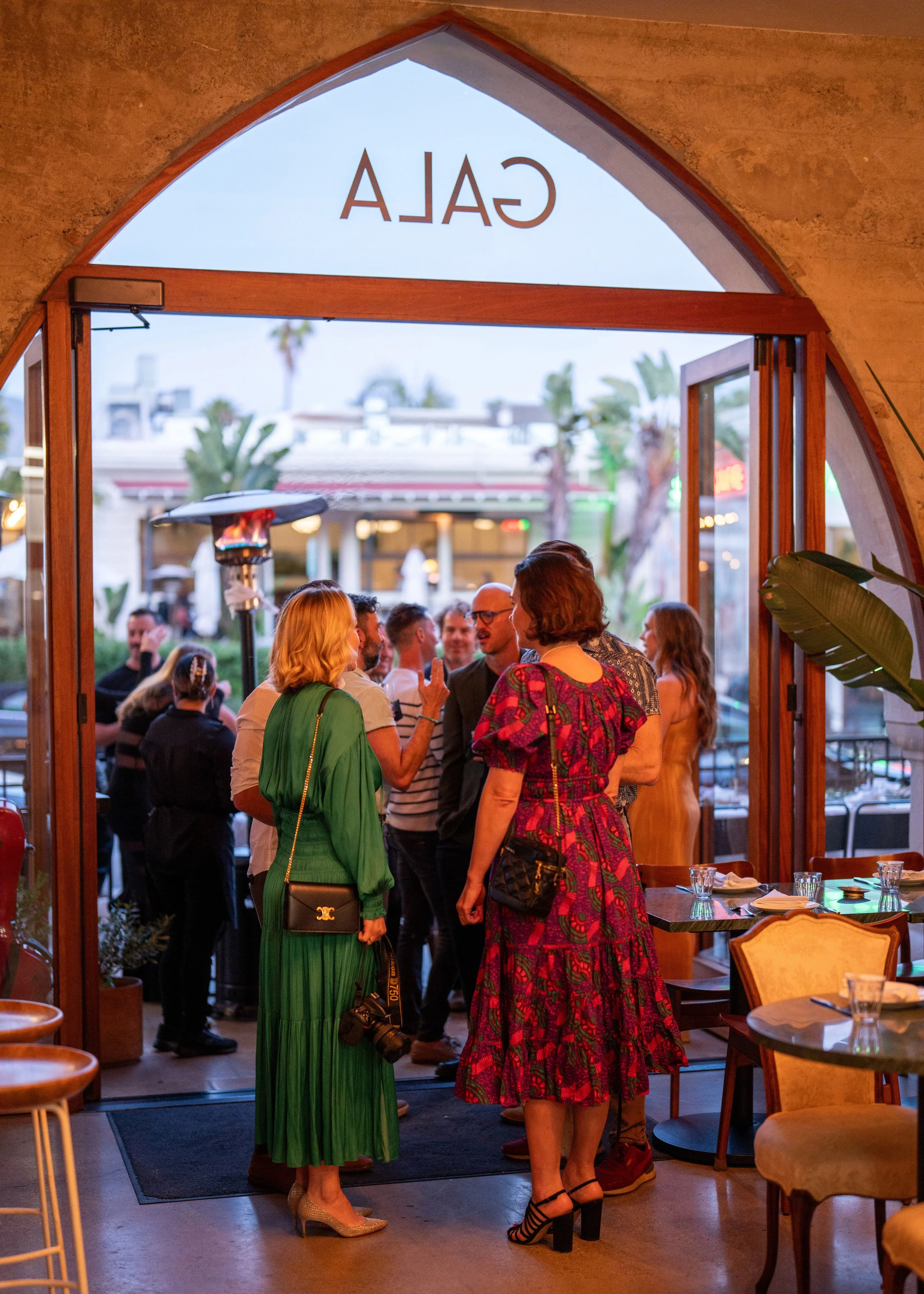 People socializing in a restaurant or cafe with large glass doors and a 'CALA' sign on the glass. Some people are standing and conversing, with tables set with glasses and napkins nearby. Outdoor scenery includes palm trees and buildings.