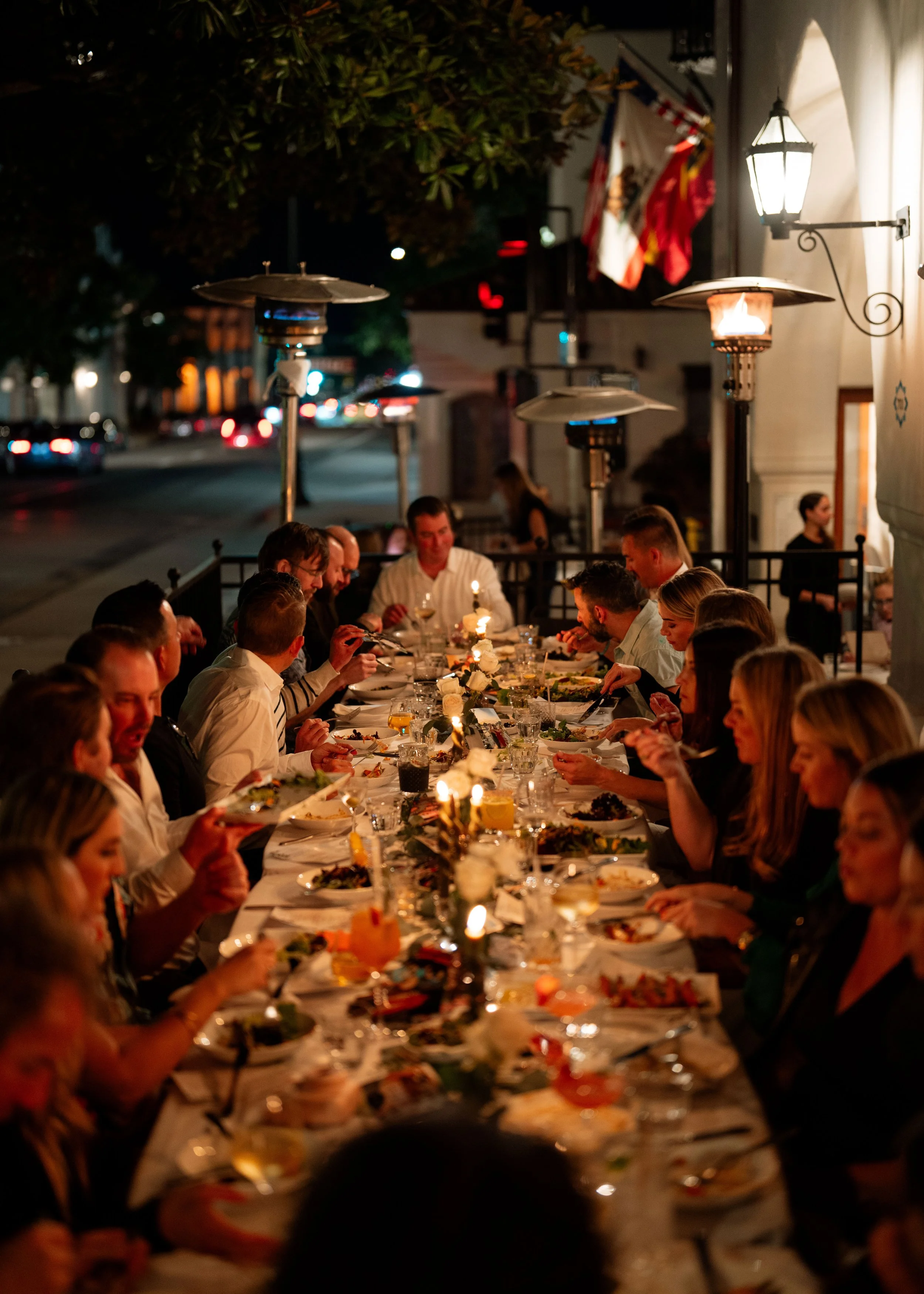 People gather for a dinner at a long table outdoors at night, illuminated by string lights and outdoor lamps, with flags hanging in the background.