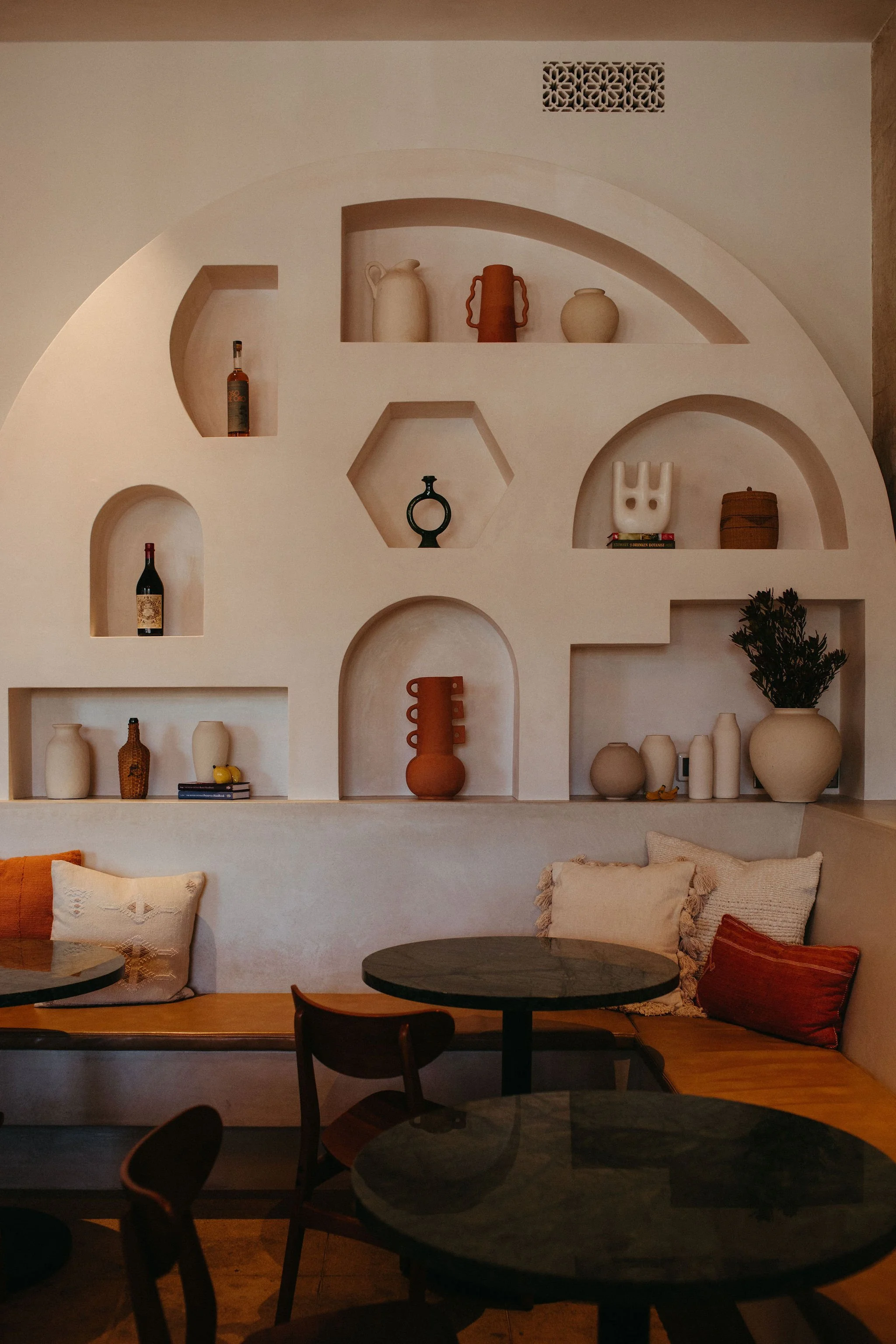 A cozy restaurant interior featuring a white wall with built-in shelves displaying decorative vases, bottles, and books. There is a seating area with cushions and a table.