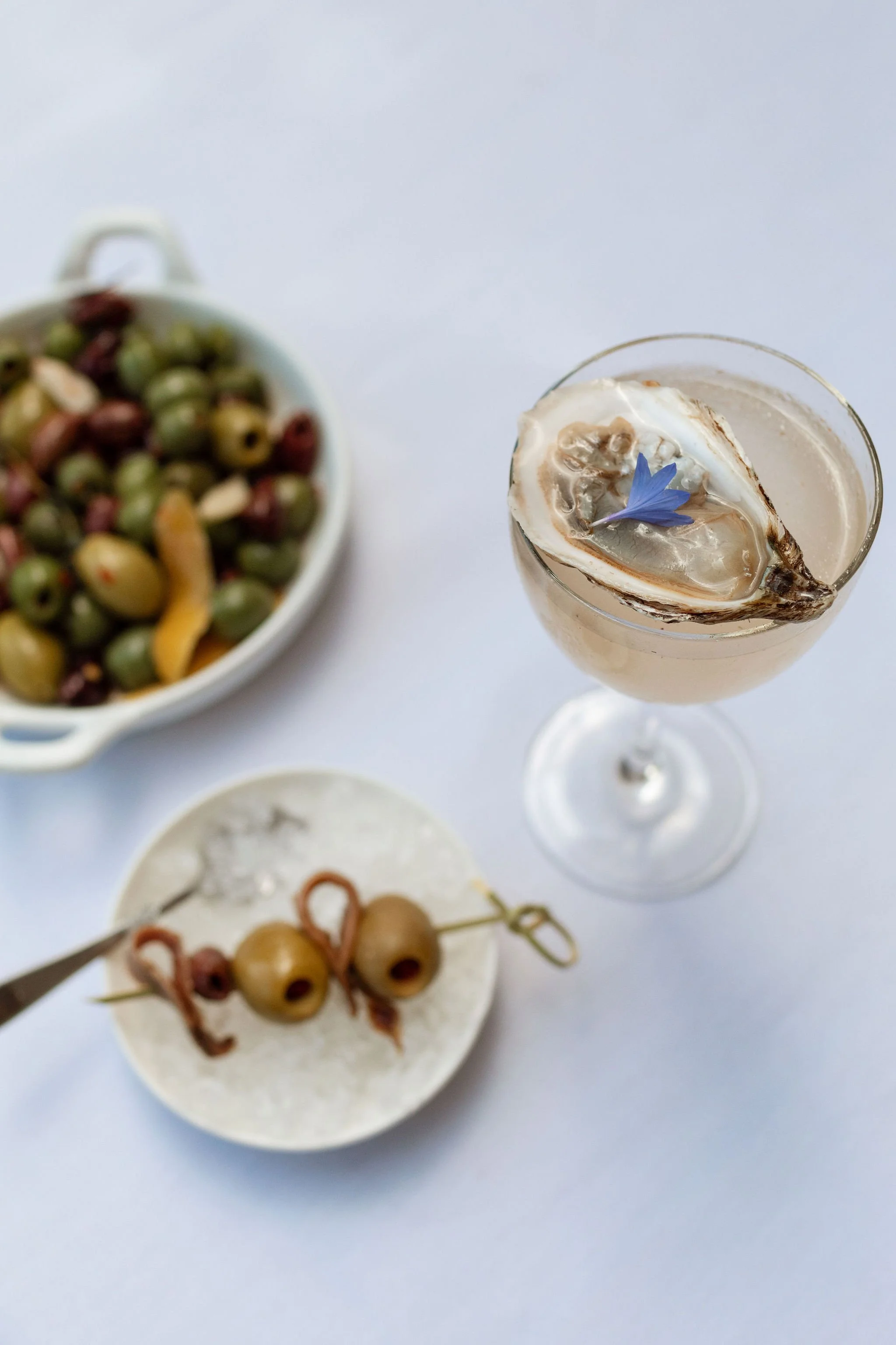 A cocktail in a tall glass garnished with a shell, a blue flower, and ice cubes. There is a bowl of mixed olives and a plate with skewered olives on a white table.