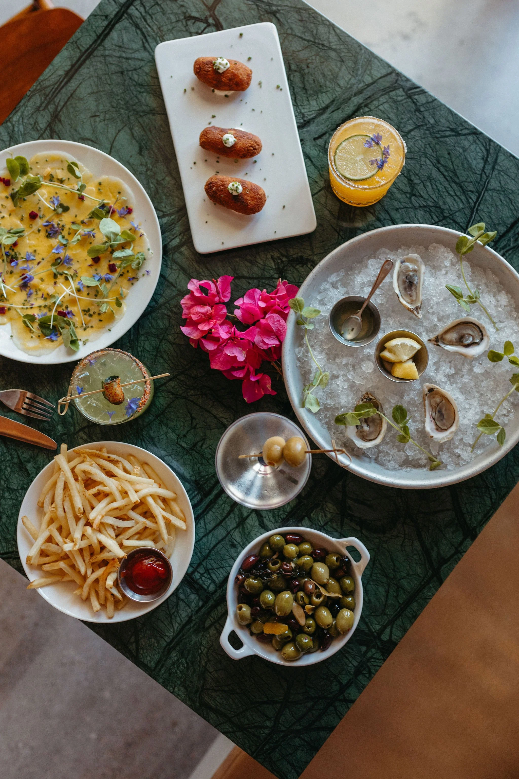 A table filled with appetizers, including oysters on ice with lemon wedges, a bowl of green olives, a plate of French fries with ketchup, a plate of ravioli garnished with microgreens and edible flowers, and a cocktail with lemon and purple flowers.
