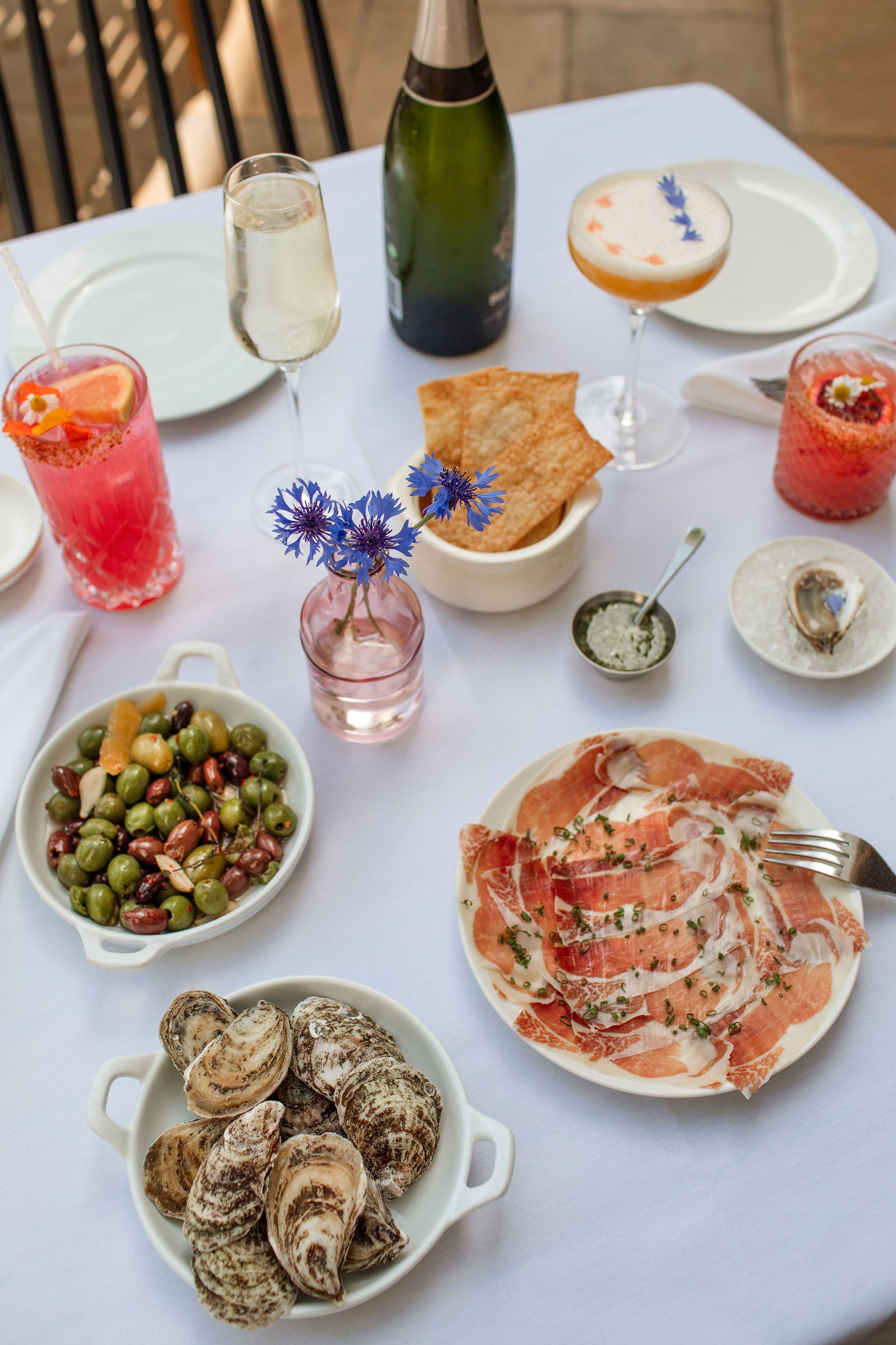 A table set for a meal with various dishes and drinks, including a bowl of oysters, a salad of mixed olives, slices of cured meats, bread, and drinks such as champagne, cocktail glasses with garnishes, and a bottle of sparkling wine.