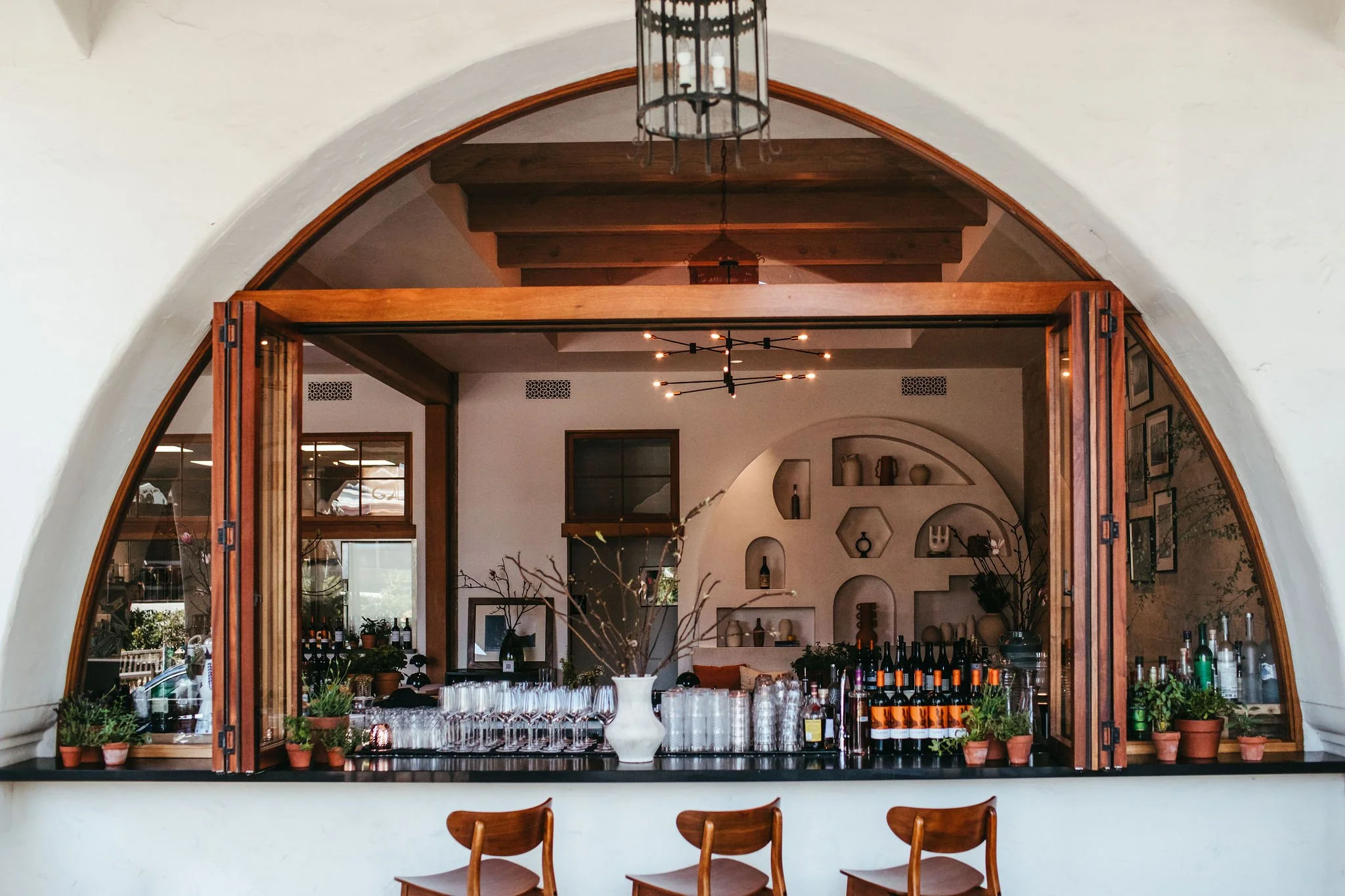 A view of a restaurant interior seen through an arched window with wooden framing. Inside, there is a bar area with bottles of wine, glassware, and potted plants on a black counter. The decor includes shelves with vases and artwork, with modern black
