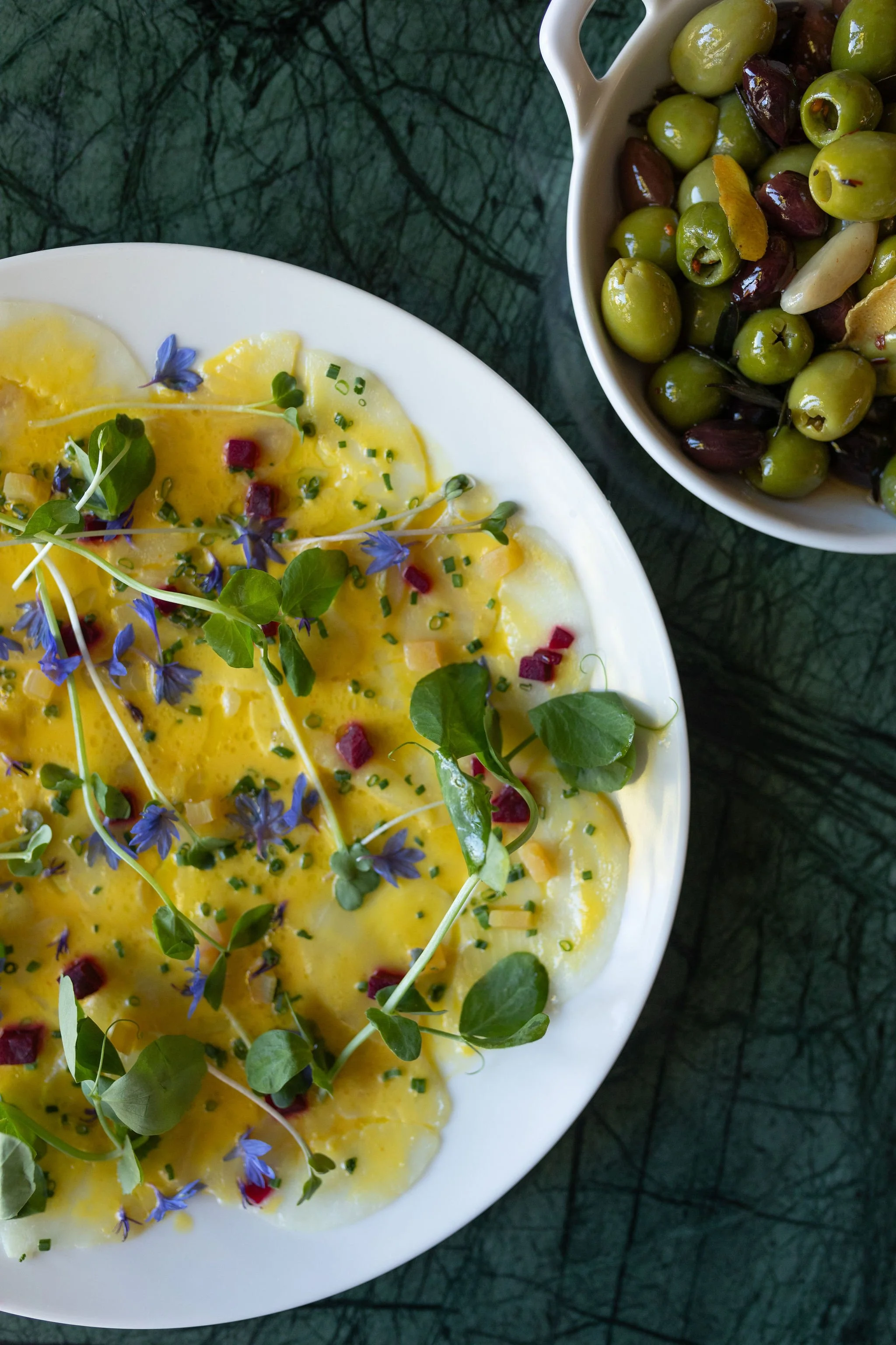 A white plate with sliced yellow eggs garnished with microgreens and edible flowers, next to a bowl of green and purple olives on a dark textured surface.