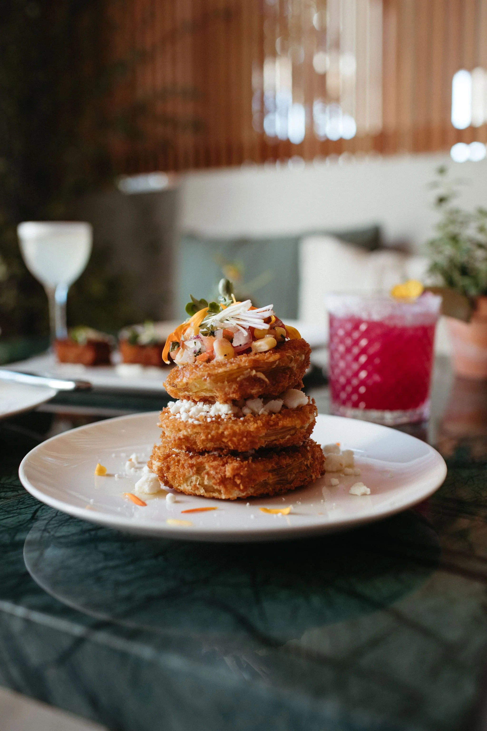 A stack of breaded onion rings topped with a colorful salad of vegetables, served on a white plate. In the background, there is a glass of pink beverage and additional plates of food, with a cozy indoor setting featuring wooden ceilings and a blurred background.