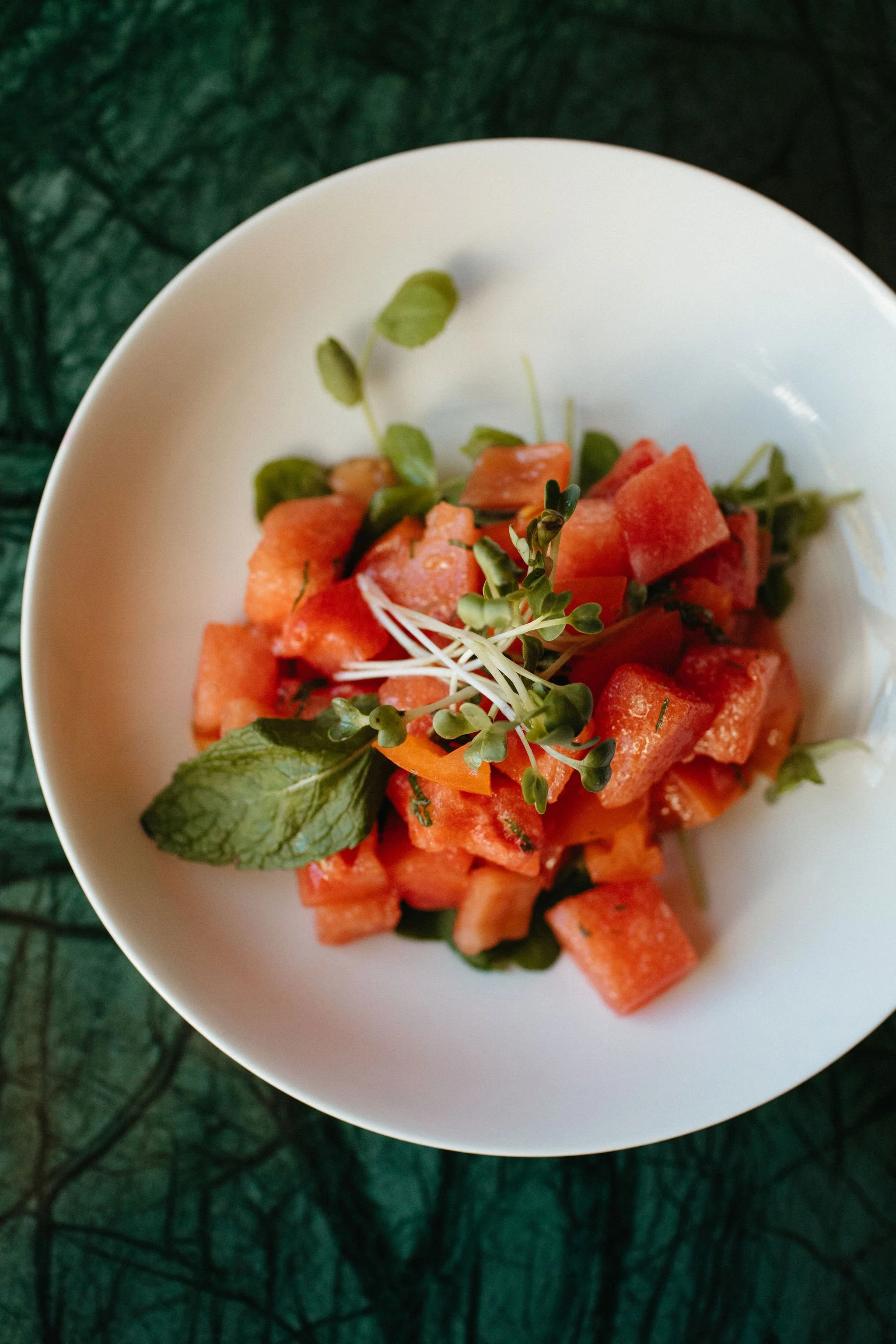 A white bowl containing chopped watermelons garnished with microgreens, with green leaves on the side, placed on a dark green textured surface.