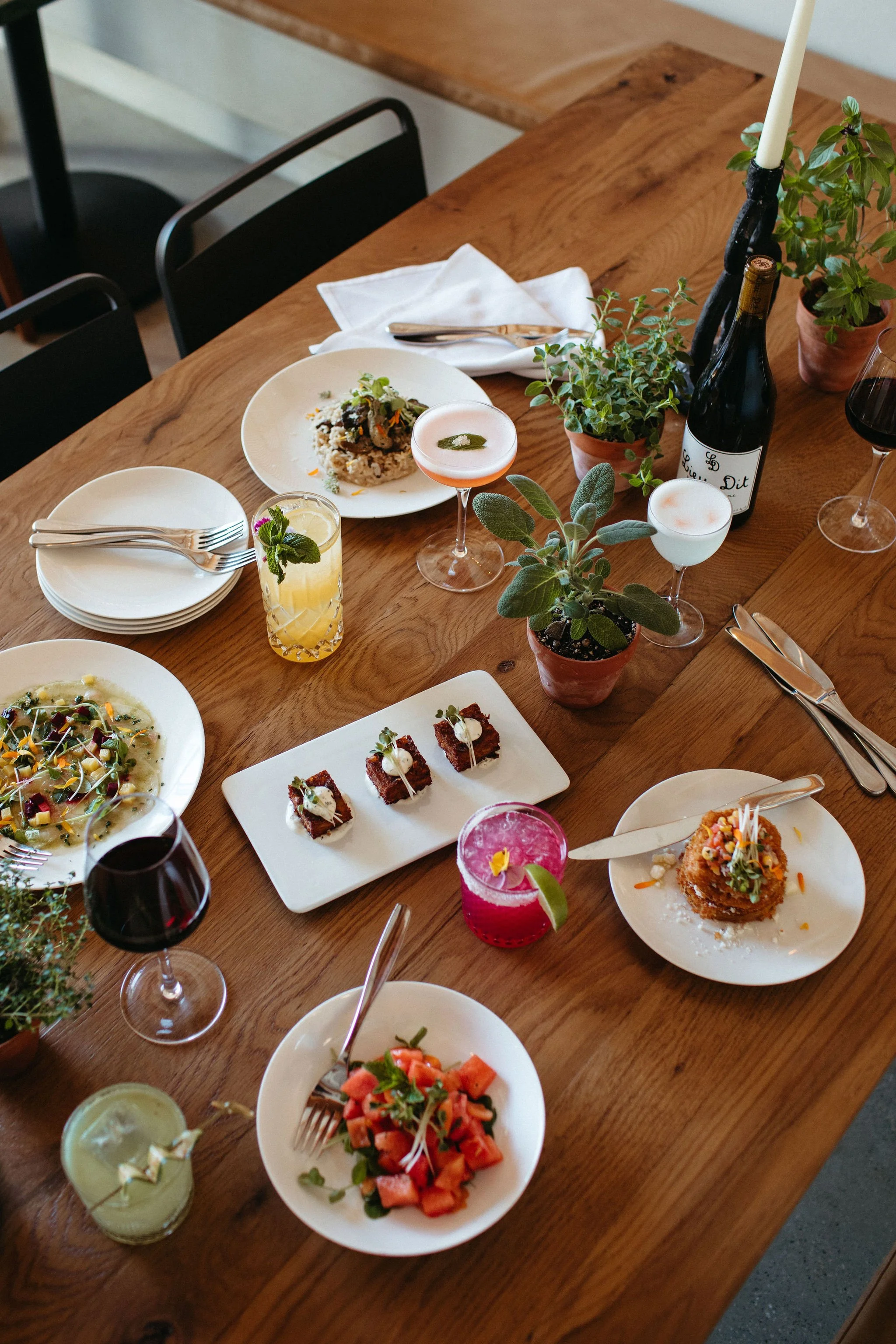 A wooden dining table set with various dishes, drinks, and plants, including a bottle of wine, cocktails, and plates of food.