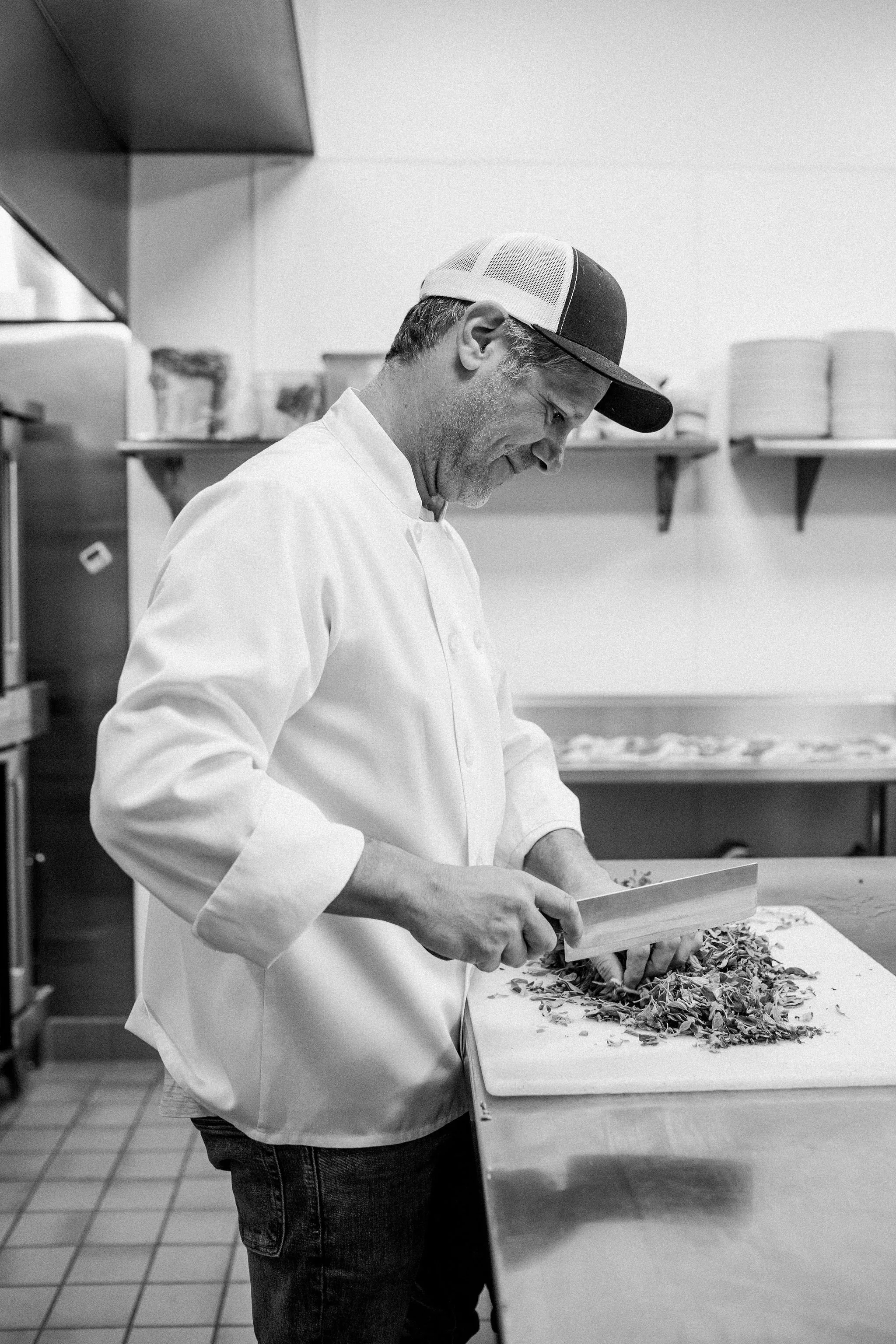 Man in chef's coat and baseball cap chopping vegetables on a cutting board in a commercial kitchen.