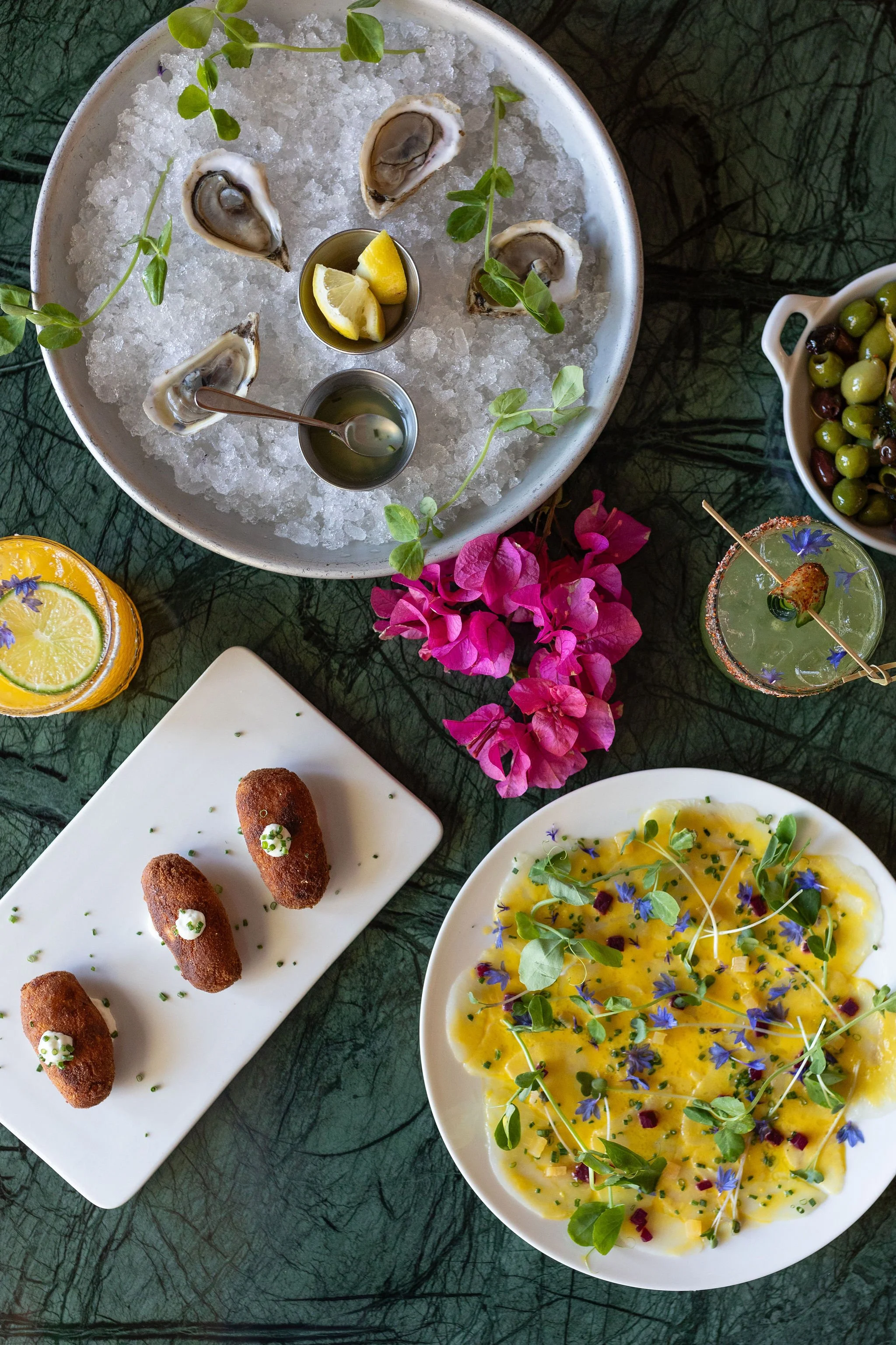 A platter of raw oysters on ice garnished with lemon wedges, accompanied by green microgreens, with a bowl of mixed olives, a cocktail with flowers, and croquettes on separate plates, all on a dark green textured surface.
