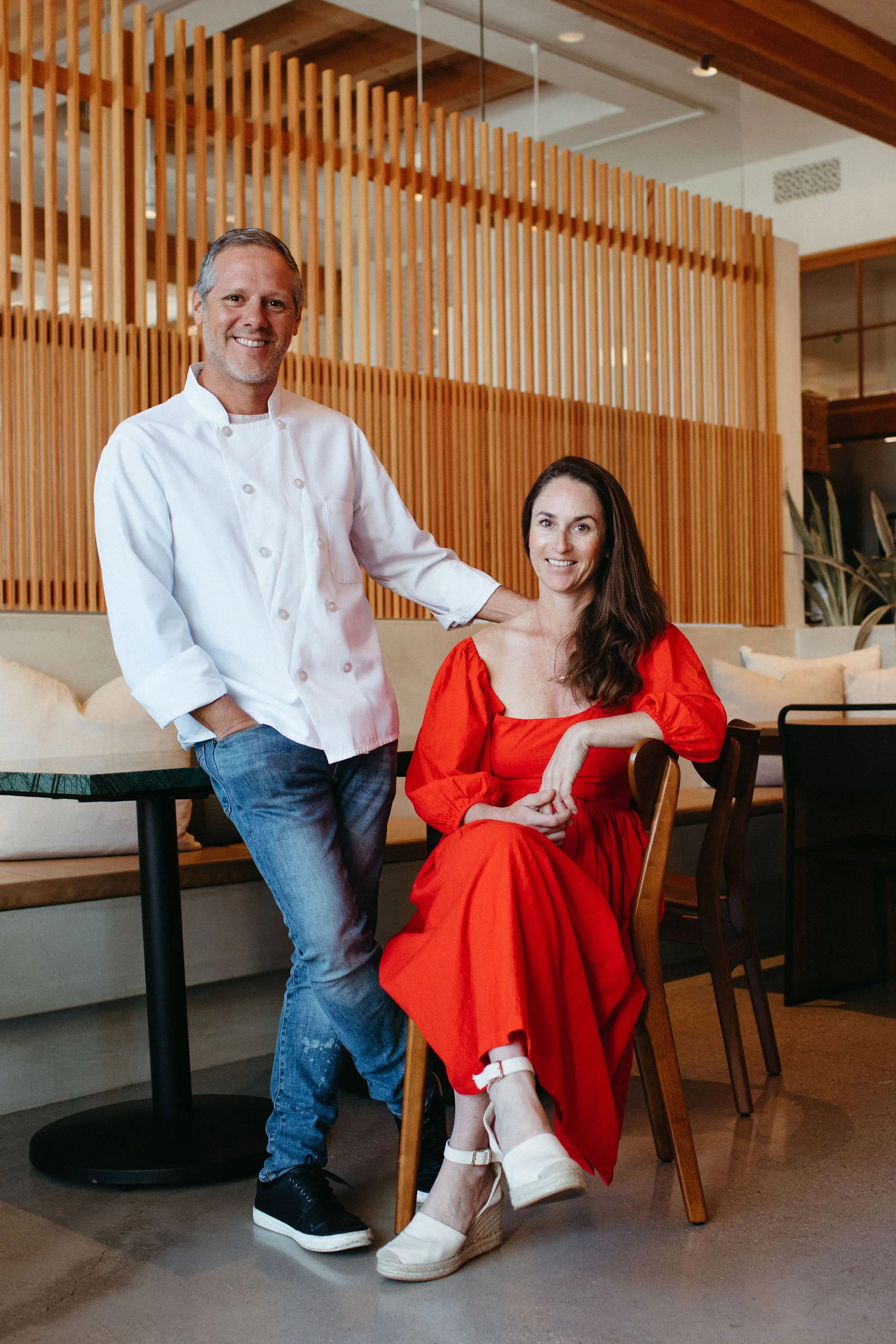 A man dressed as a chef standing next to a woman sitting in a dining area with wooden decor behind them. The woman is wearing a red dress and white shoes, and is smiling at the camera. The man is smiling, wearing a white chef's coat, and has his hand on the woman's shoulder.