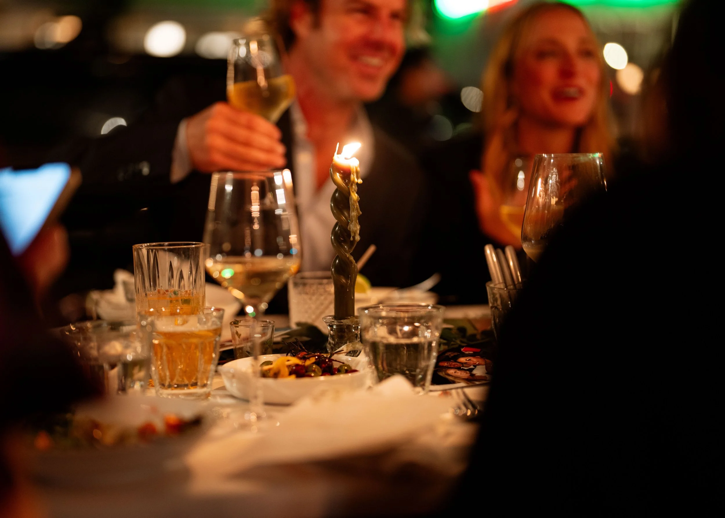 People celebrating at a dinner party with drinks, a lit candle, and food on the table in a festive setting.