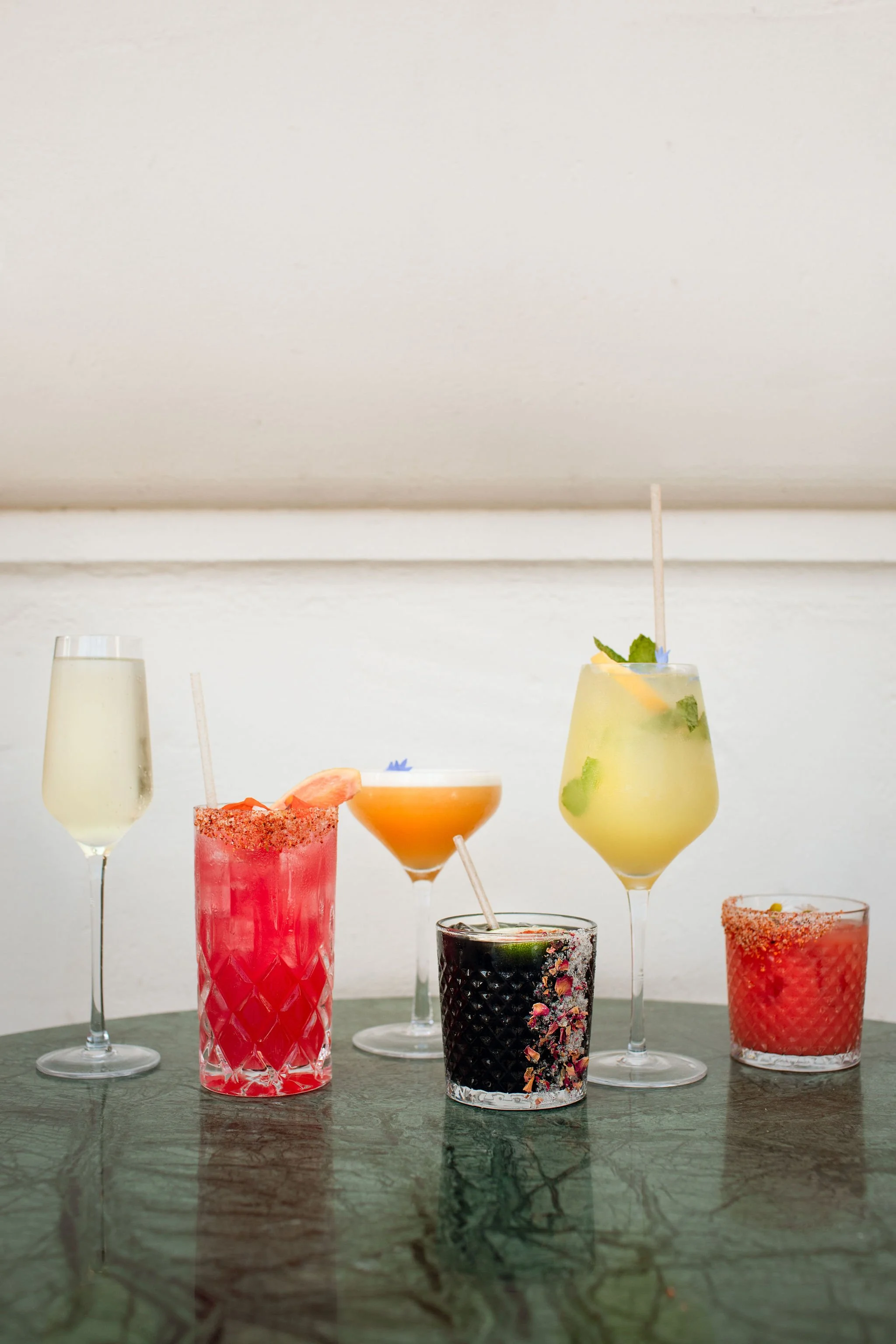A variety of colorful cocktails on a marble table, including a yellow drink garnished with mint, a black drink with colorful sprinkles, a pink drink with a salted rim, an orange drink, a clear drink with a slice of fruit, and a red drink with a salte